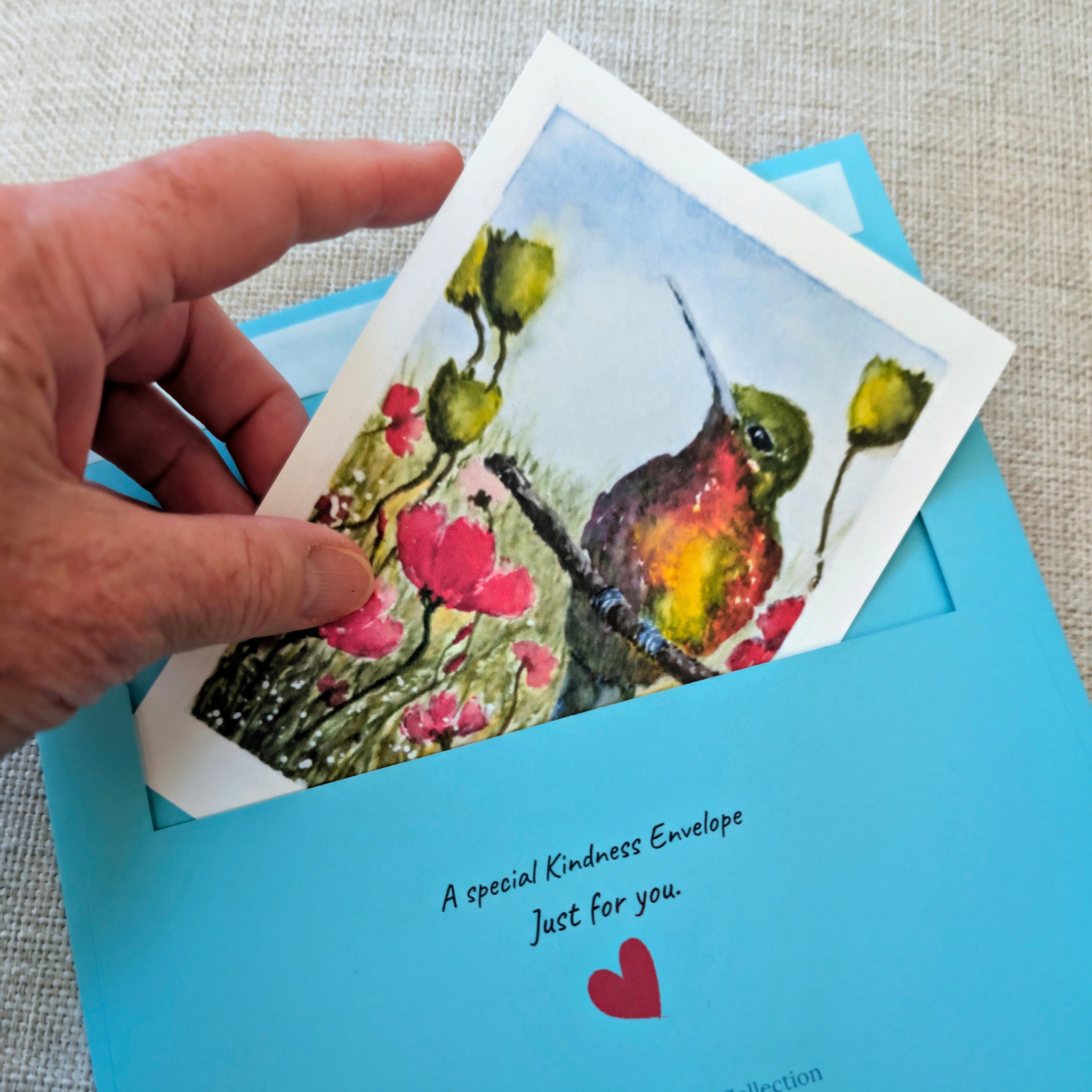 Hand holding an original hummingbird watercolor above a blue Kindness Envelope, showing the handmade nature of each mailing.