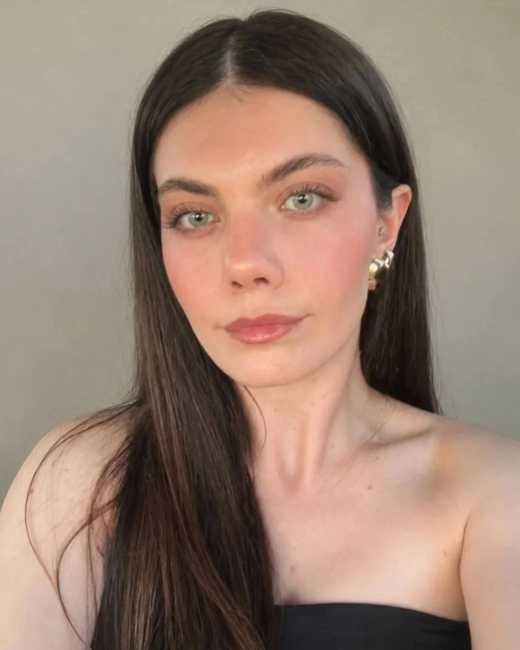 A woman with long dark hair, light skin, and green eyes looking at the camera, wearing earrings and a black strapless top.