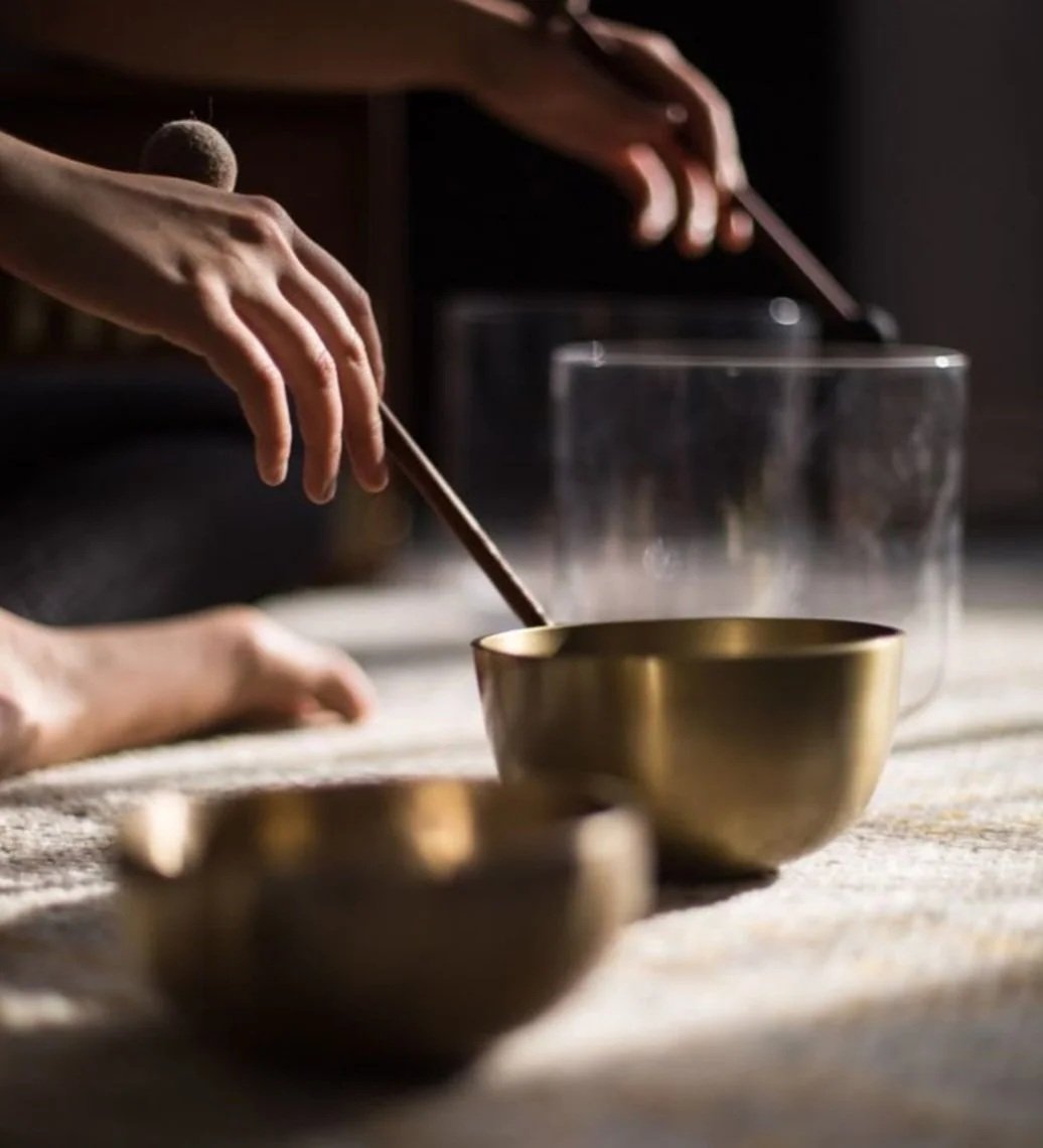 Close-up of hands striking a singing bowl with a wooden mallet, with another partly visible singing bowl in the foreground and a glass container in the background.