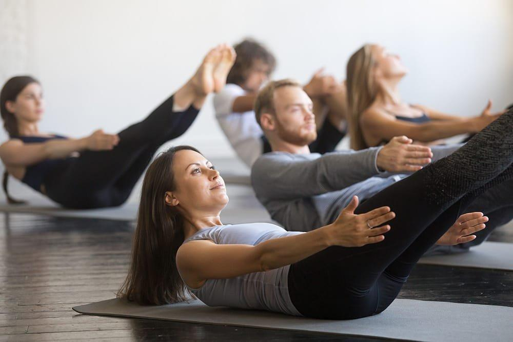 Group of men and women doing yoga on mats in a studio, lying on their backs with legs and arms raised, practicing core and balance exercises.