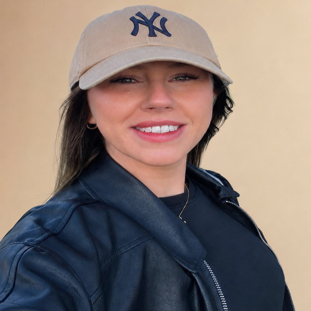 A woman with shoulder-length brown hair, wearing a beige Yankees cap, a black leather jacket, and a black top, smiling against a beige background.