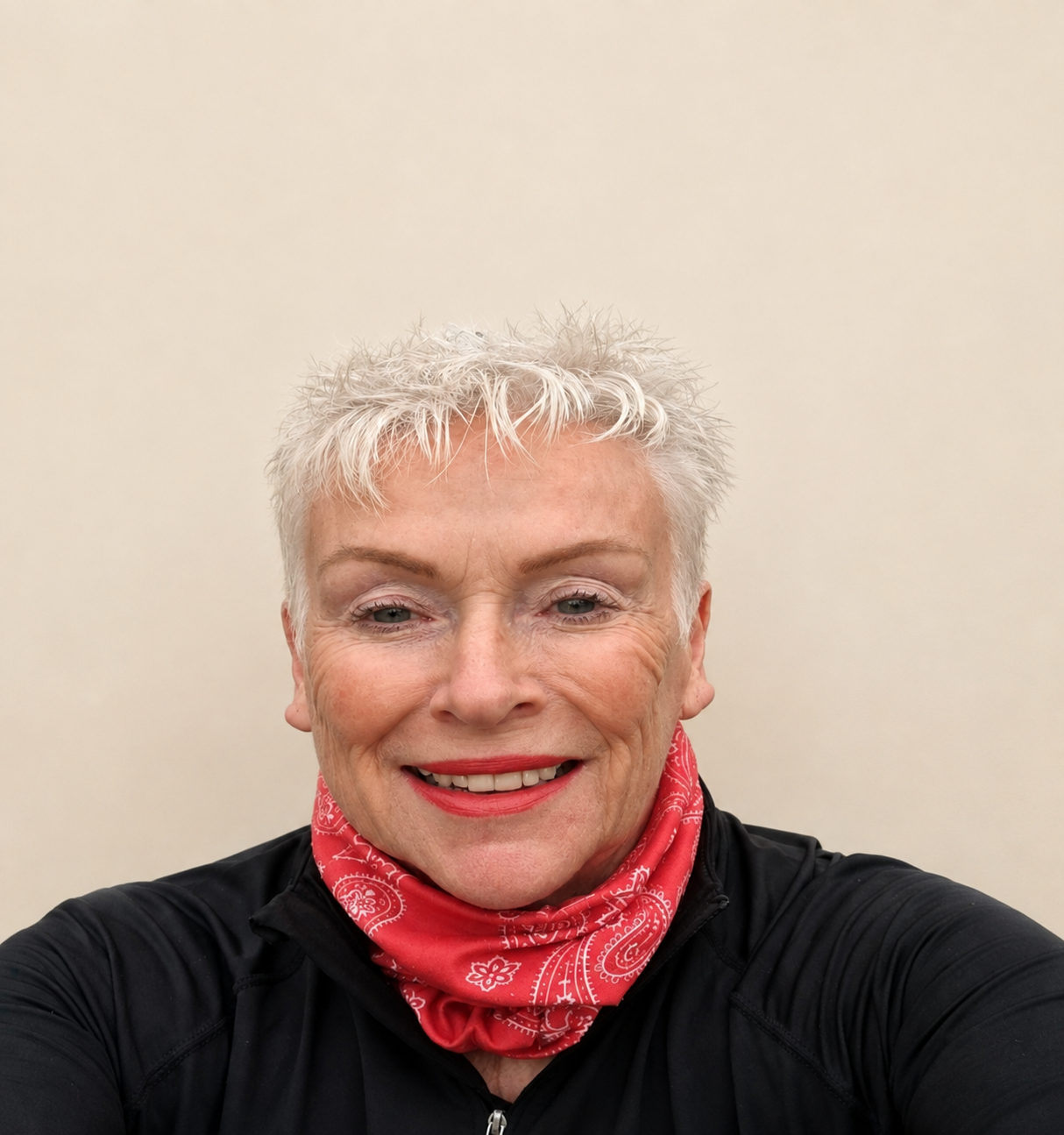 A woman with short, spiky, gray hair, wearing a red bandana around her neck and a black jacket, smiling in front of a plain light-colored wall.
