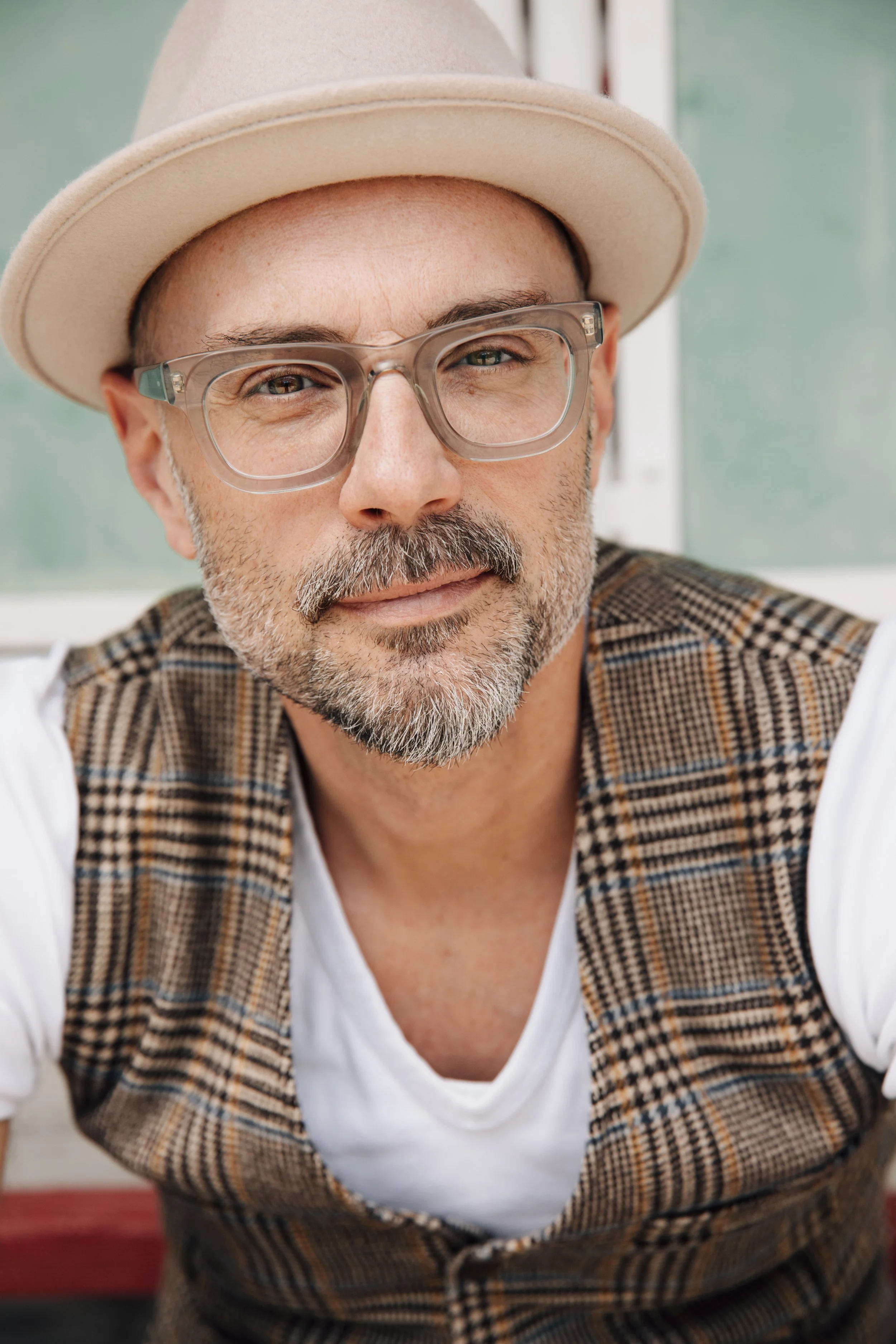 Close-up of a middle-aged man with glasses, wearing a beige fedora, plaid vest, and white shirt, smiling and looking at the camera.