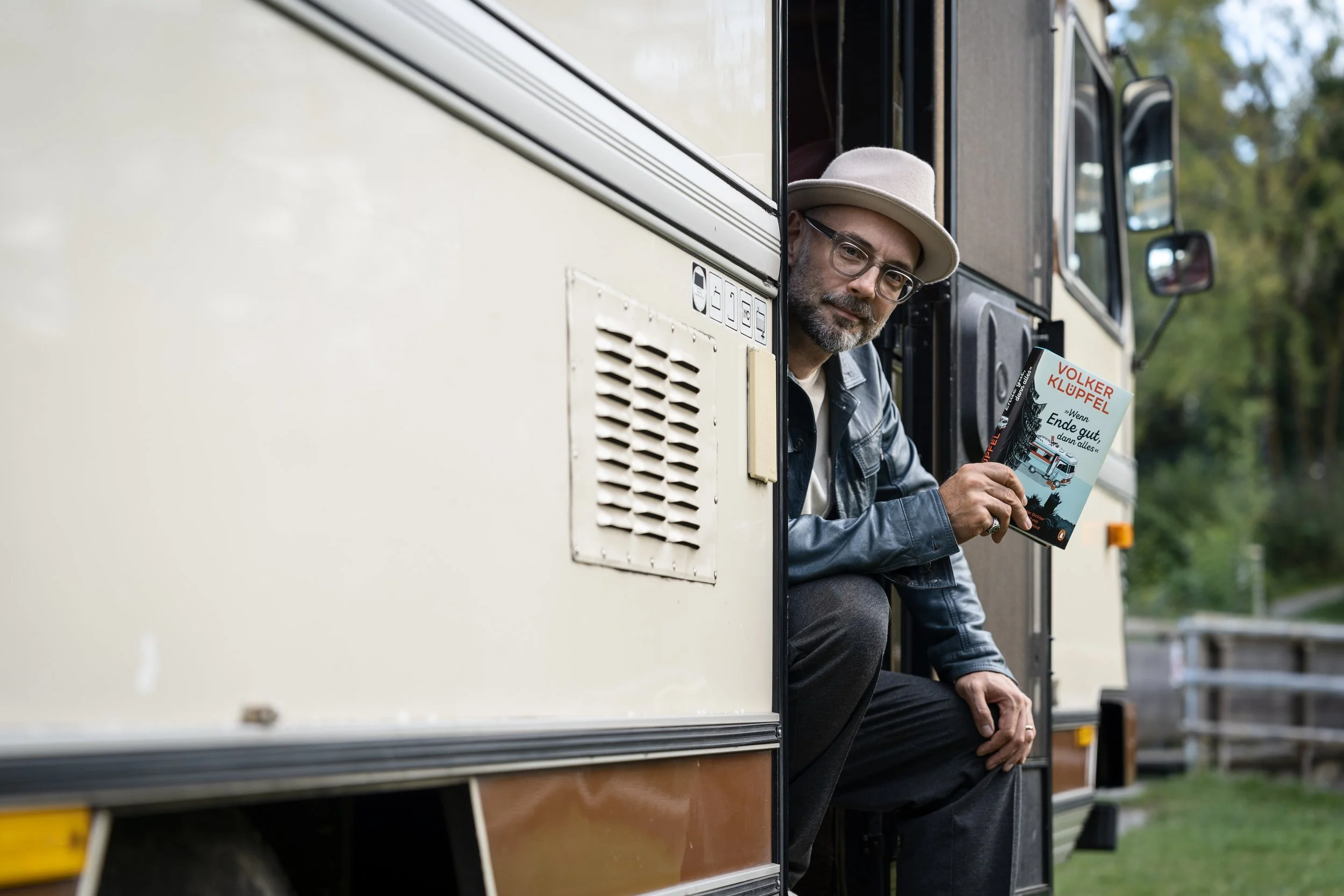 A man with glasses, a beige hat, and a leather jacket sitting in a trailer, holding a book titled "Wenn Ende gut" by Volker Klüpel, with trees in the background.