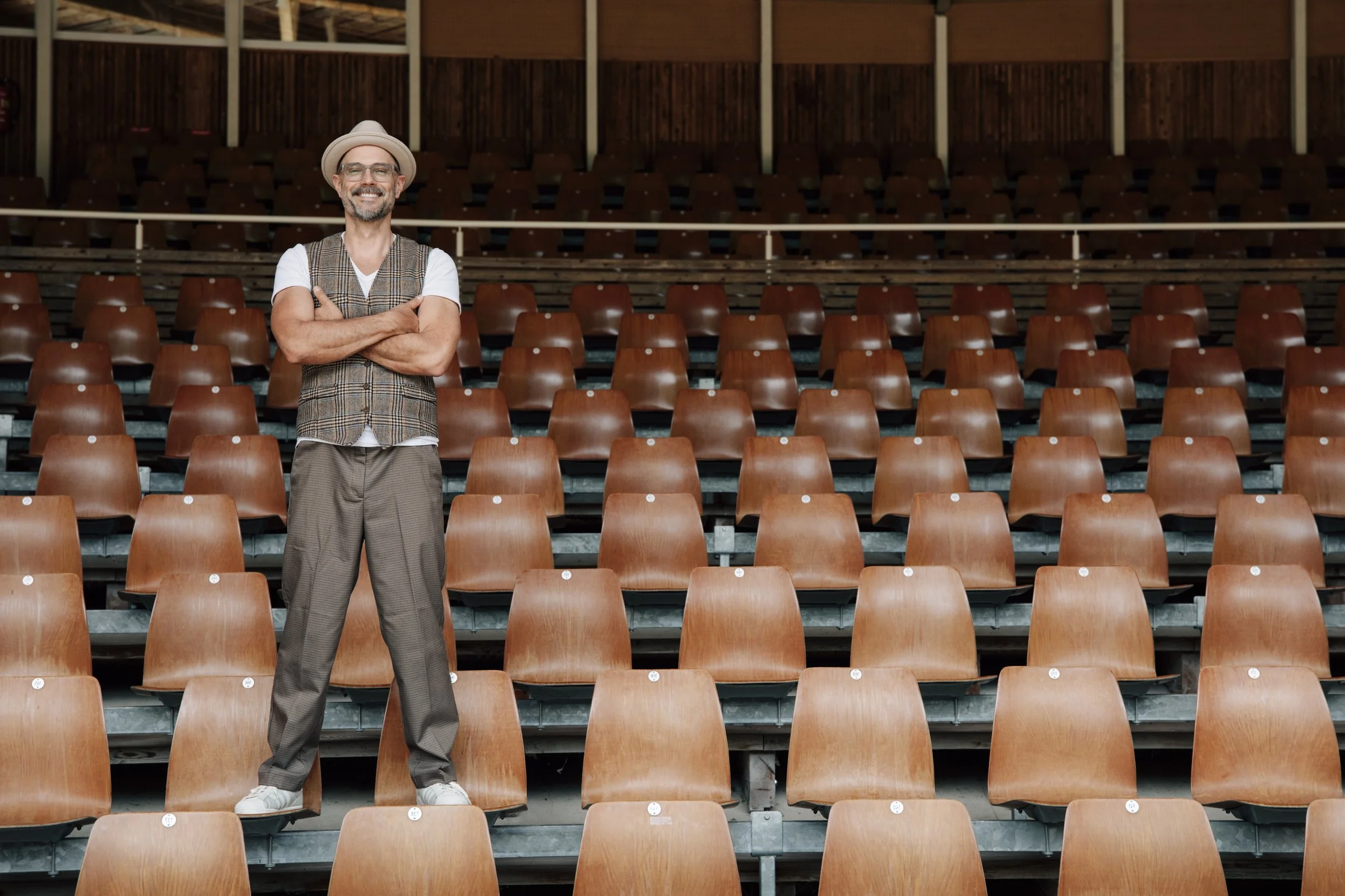 A man smiling and standing with arms crossed in an empty auditorium with wooden seats and a stage.