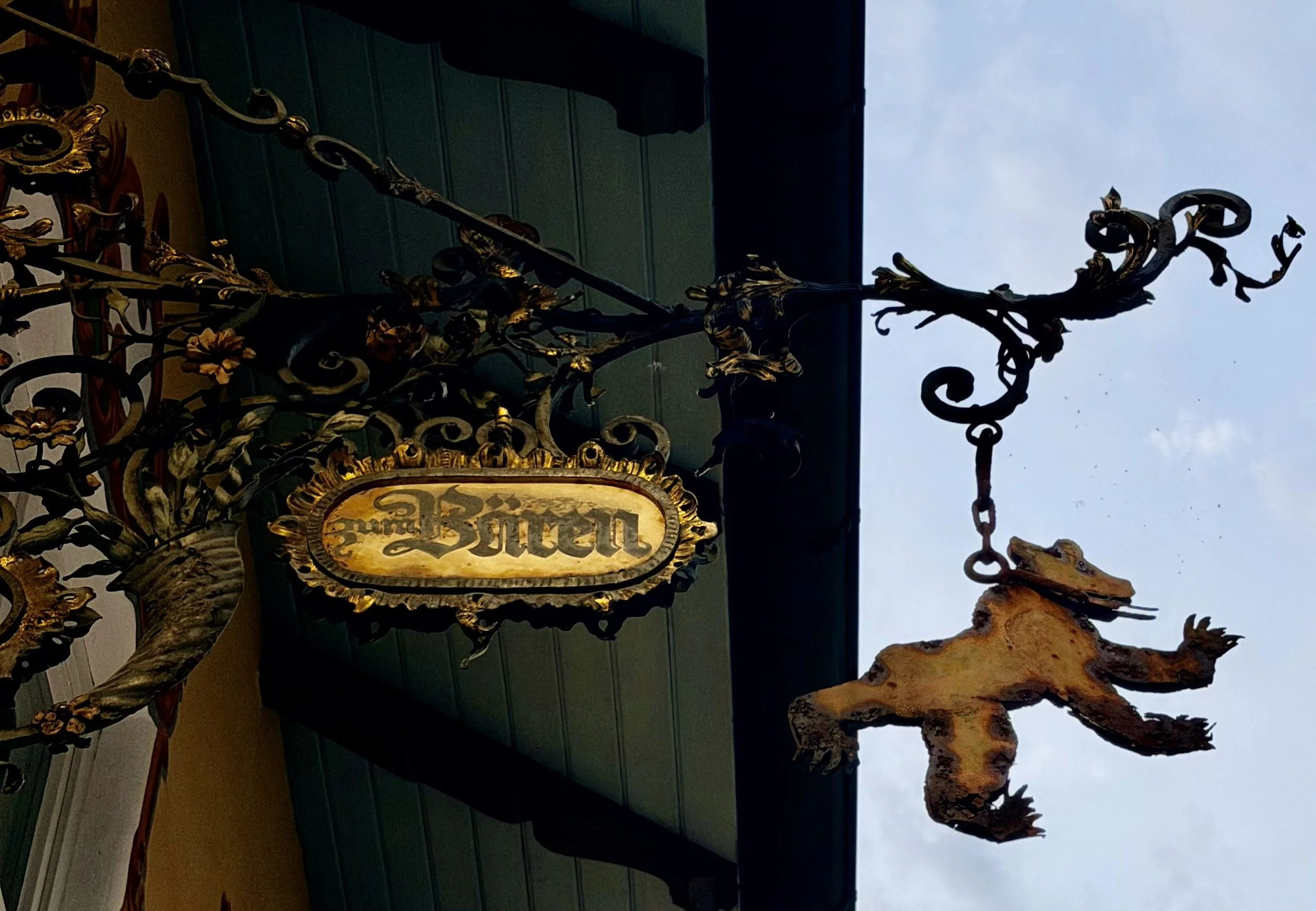 An ornate black wrought iron sign with gold accents, hanging from a decorative metal bracket, reads 'Buken'. The sign is mounted on the exterior of a building with dark teal horizontal siding. The sky above is overcast.