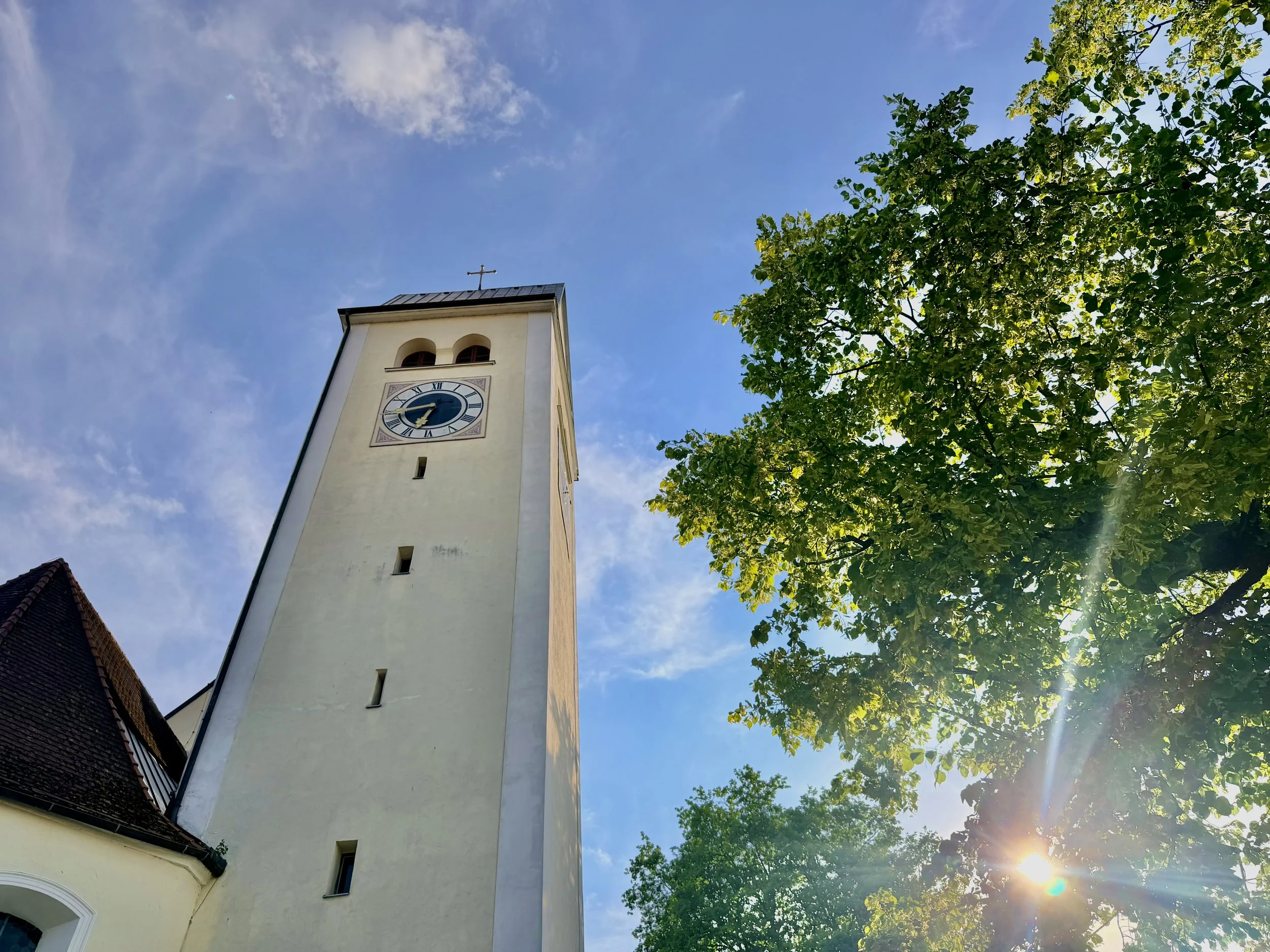 View of a church tower with a clock face from below, against a blue sky with some clouds and green trees. The sun is shining, creating a lens flare.