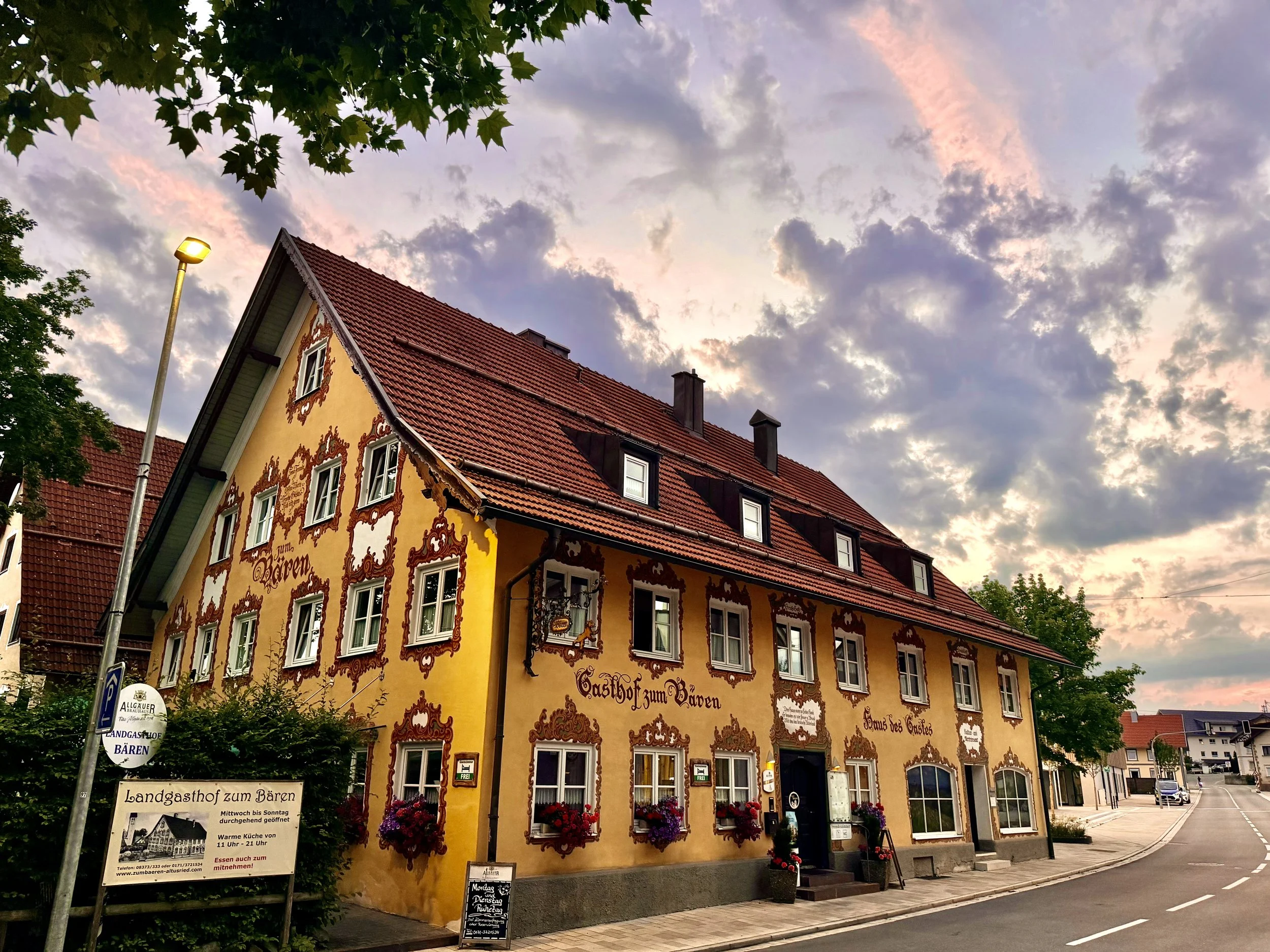 A colorful building with traditional Bavarian architecture during sunset, featuring a red-tiled roof, ornate window frames, and floral decorations, with a sign that reads 'Landgasthof zum Bären' in German.