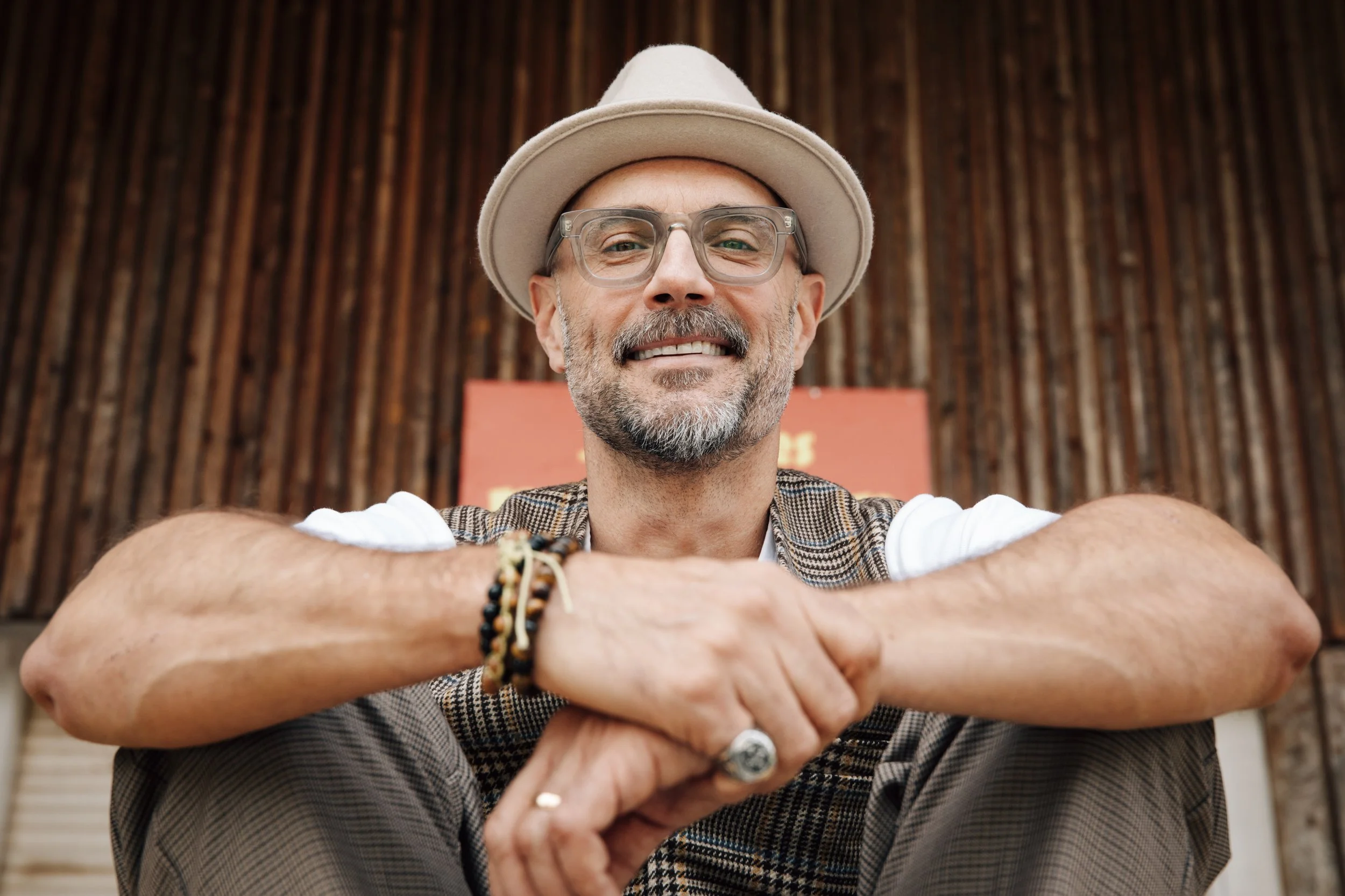 Close-up of a smiling man wearing glasses and a beige hat, sitting cross-legged with arms resting on his knees, against a wooden background.