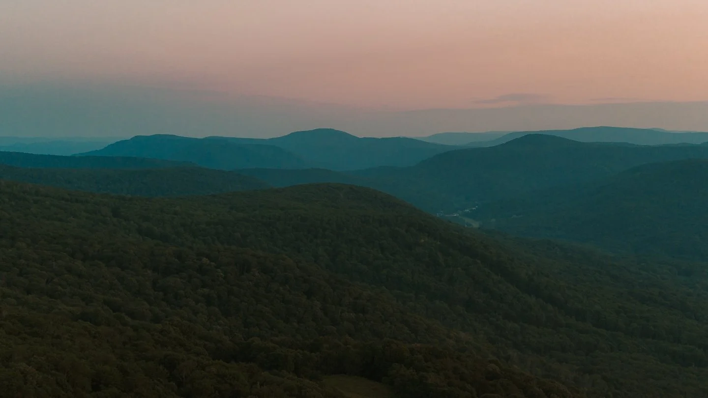 Distant mountain range at dusk with a gradient pink and blue sky, rolling green hills in the foreground.