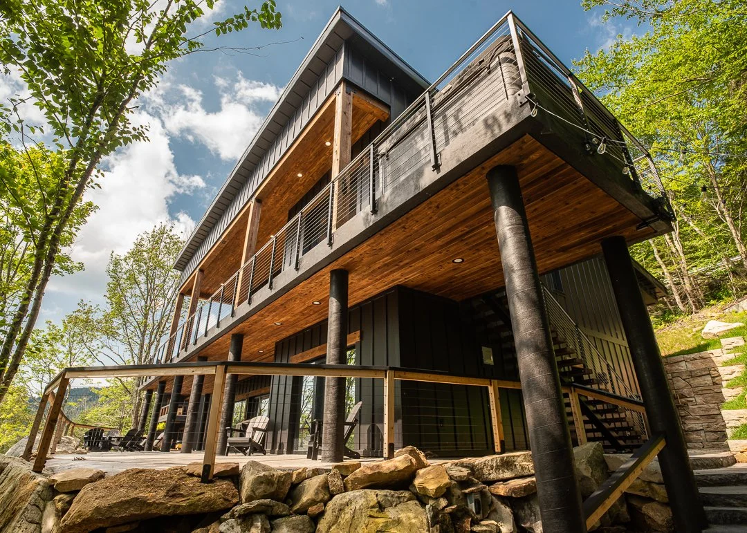 Modern two-story house with black exterior and wooden accents, elevated on stilts, with a spacious outdoor deck and railing, surrounded by trees and rocks, under a partly cloudy sky.
