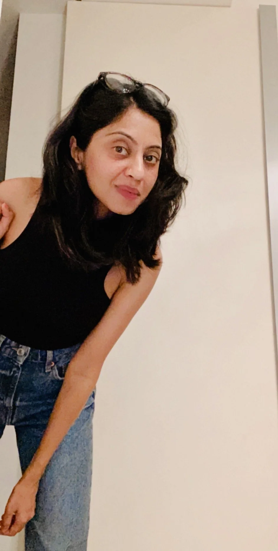 Woman with long dark hair and glasses on her head wearing a black sleeveless top and blue jeans