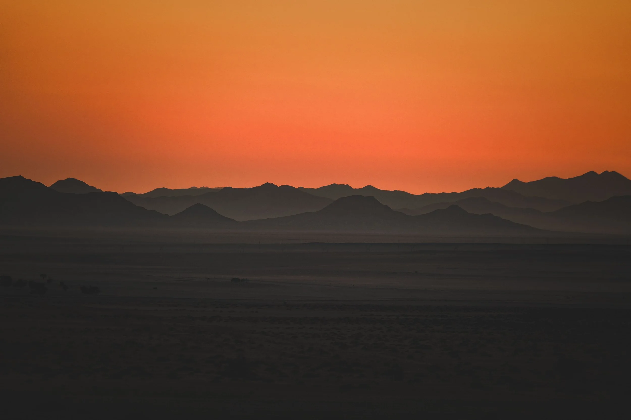Silhouetted mountain range beneath a vivid orange sunset over a dark desert landscape
