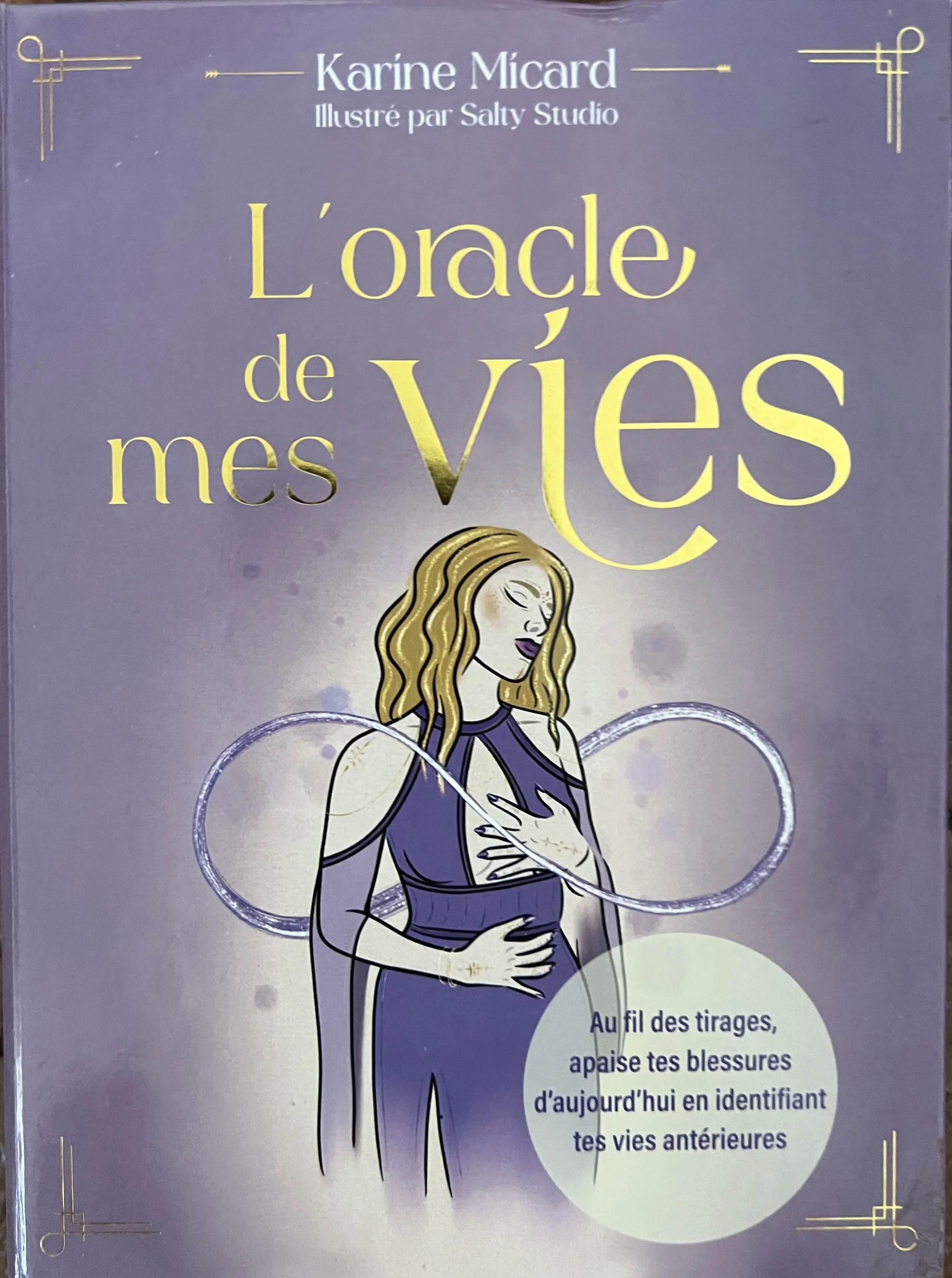 Couverture d'un livre illustré intitulé 'L'oracle de mes vies' par Karine Micard, illustré par Salty Studio. Illustration d'une femme avec long cheveux ondulés dorés, portant une robe violet foncé, entourée de cercles lumineux.