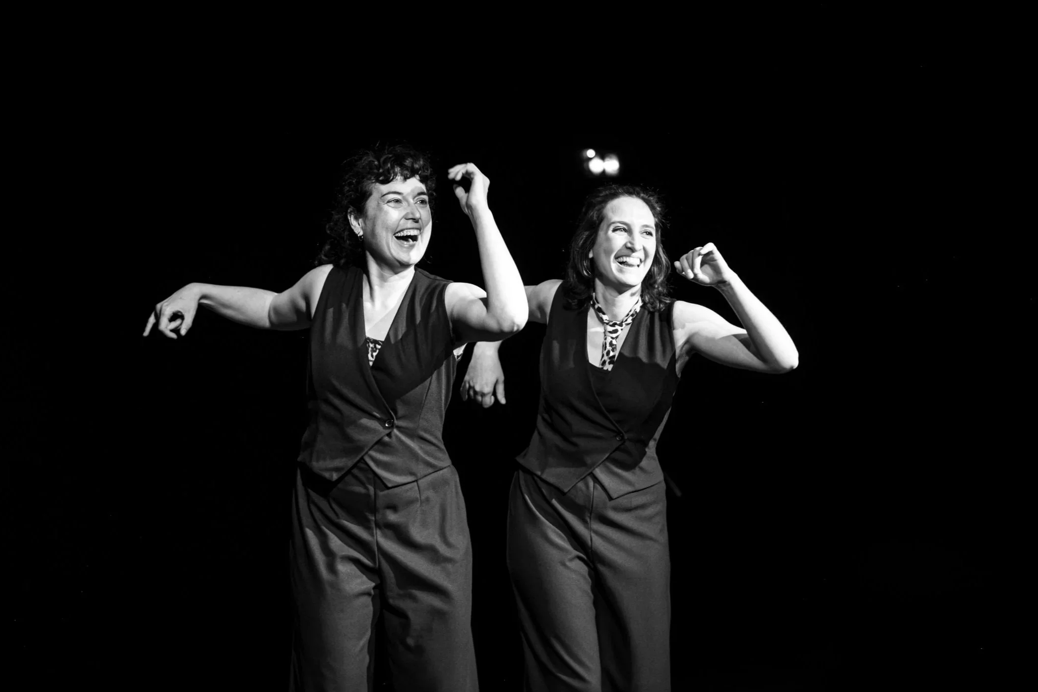 Deux femmes souriantes en costumes sombres, se tenant par la main, dans un décor noir, en train de danser ou de faire une performance sur scène.