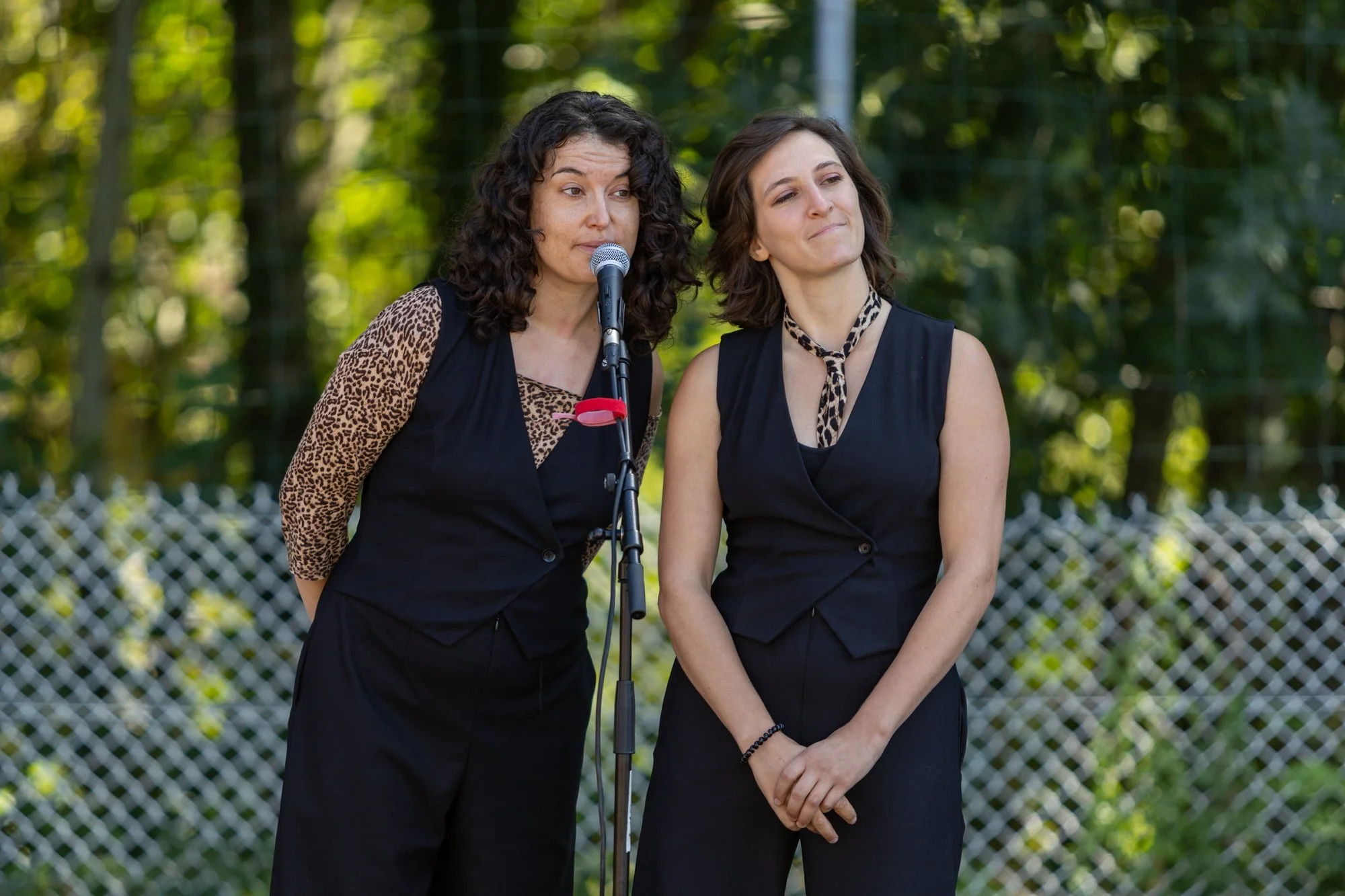 Deux femmes en noir, l'une avec une veste à motif léopard, parlent lors d'une interview ou d'un discours en plein air, avec un fond de clôture en métal et de la végétation.