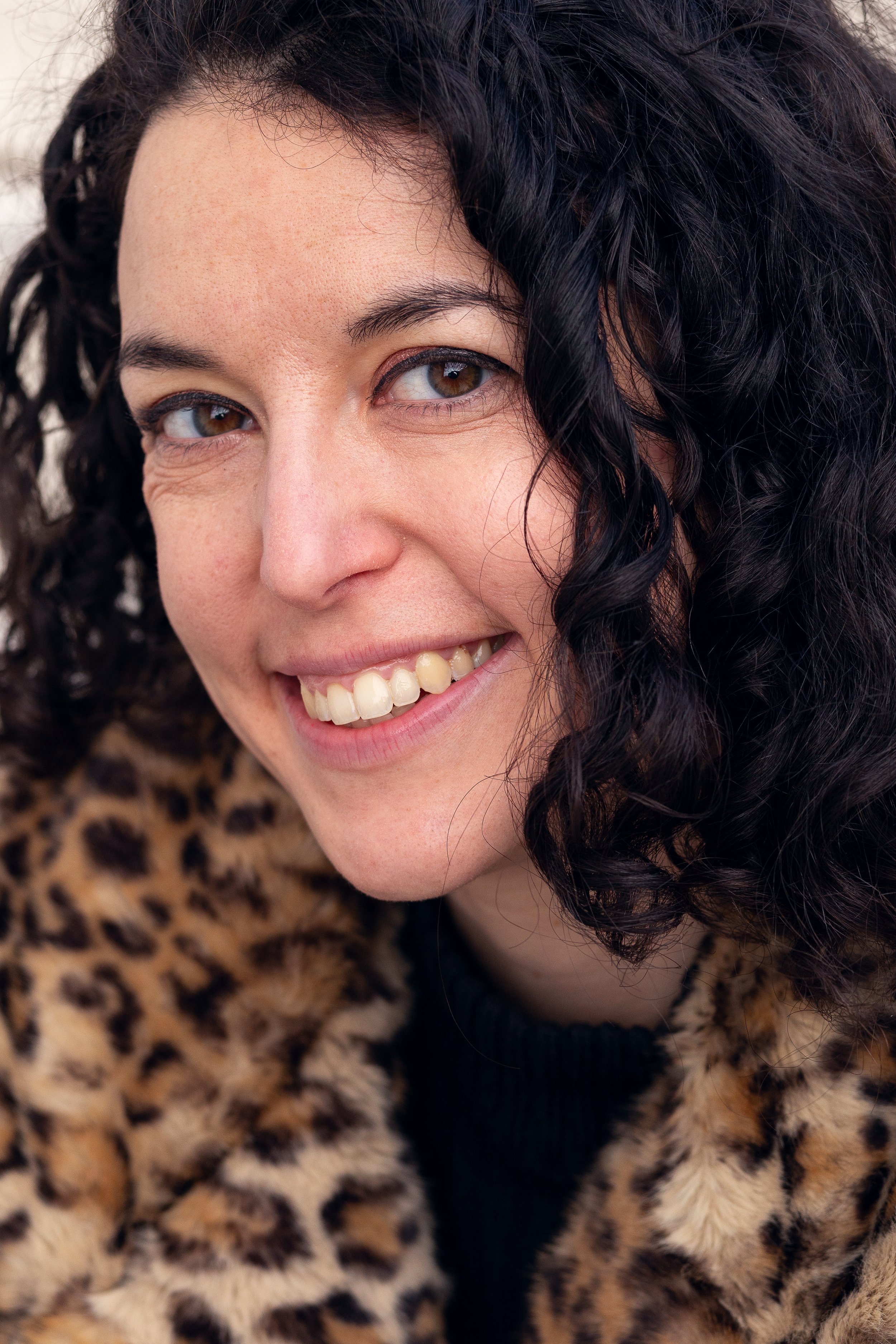 Une femme souriante avec des cheveux bouclés noirs, portant un manteau à imprimé léopard.