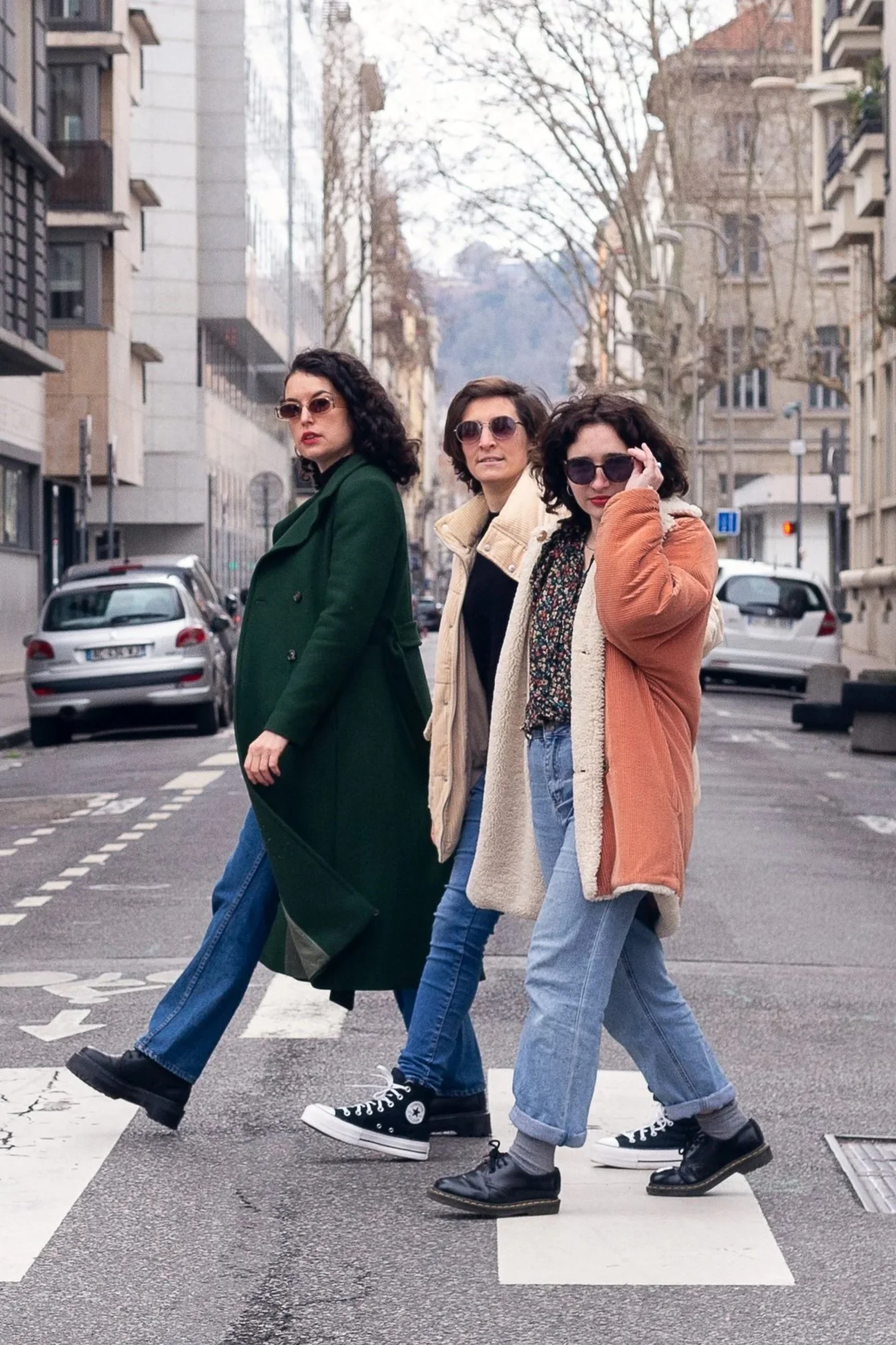 Trois femmes portent des lunettes de soleil et traversent un passage piéton dans une rue urbaine avec des bâtiments modernes. Elles ont des vêtements décontractés avec des manteaux colorés.