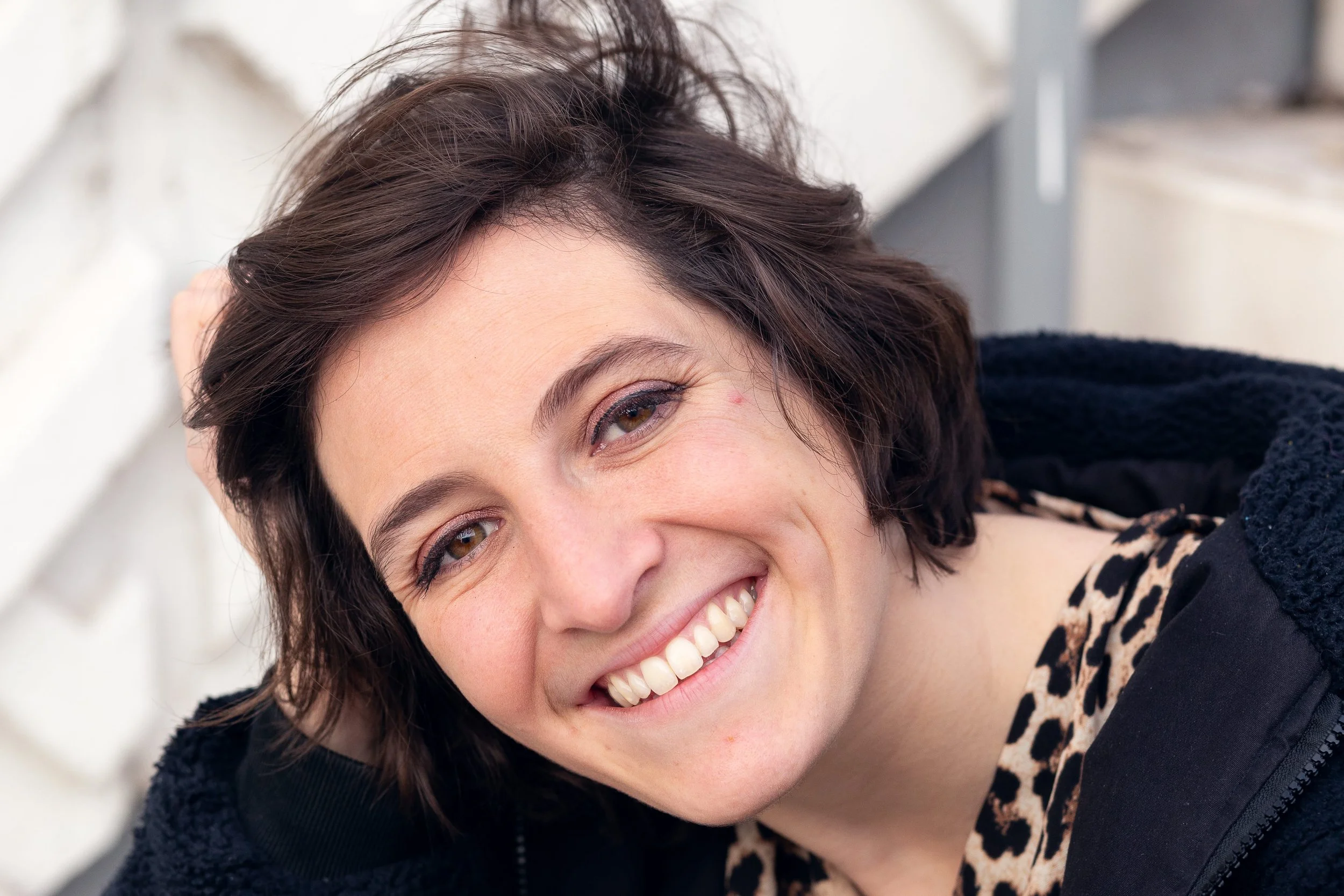 Une femme souriante avec des cheveux bruns courts repose sur un canapé ou un lit, portant un vêtement à motif animalier.