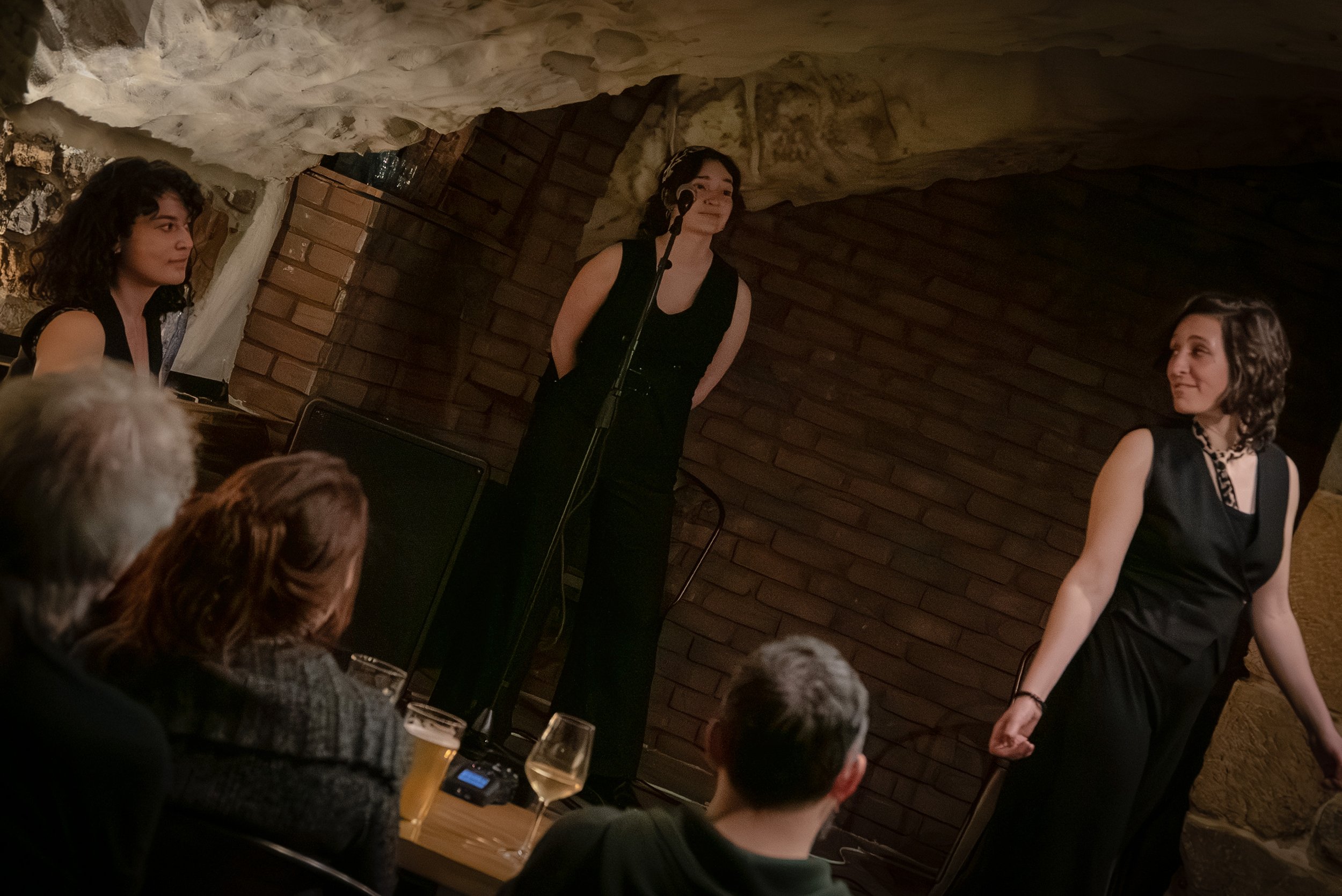 Groupe de femmes qui chante lors d'une performance dans un espace avec mur en briques et plafond en roches, certains spectateurs visibles à l'avant.
