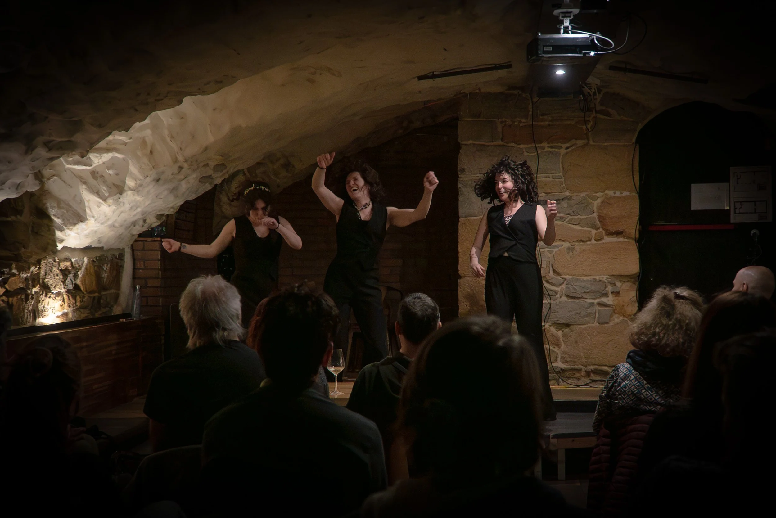 Trois femmes qui dansent ou chantent sur scène dans une cave ou un sous-sol, avec un public regardant. La scène est éclairée, avec des murs en pierre et un plafond voûté.