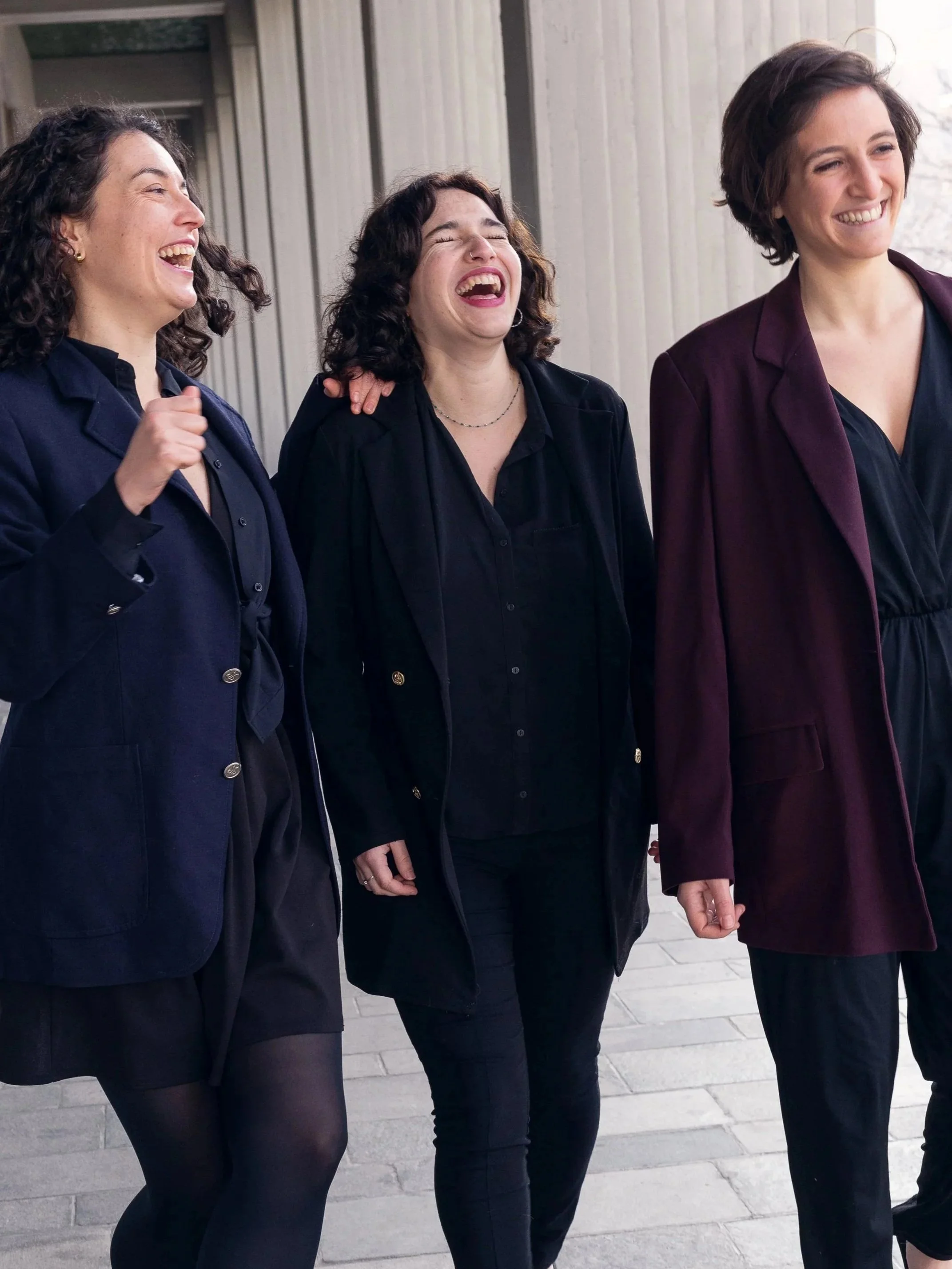 Trois femmes debout côte à côte dans une rue, souriantes et riant ensemble, portant des vêtements noirs et colorés.