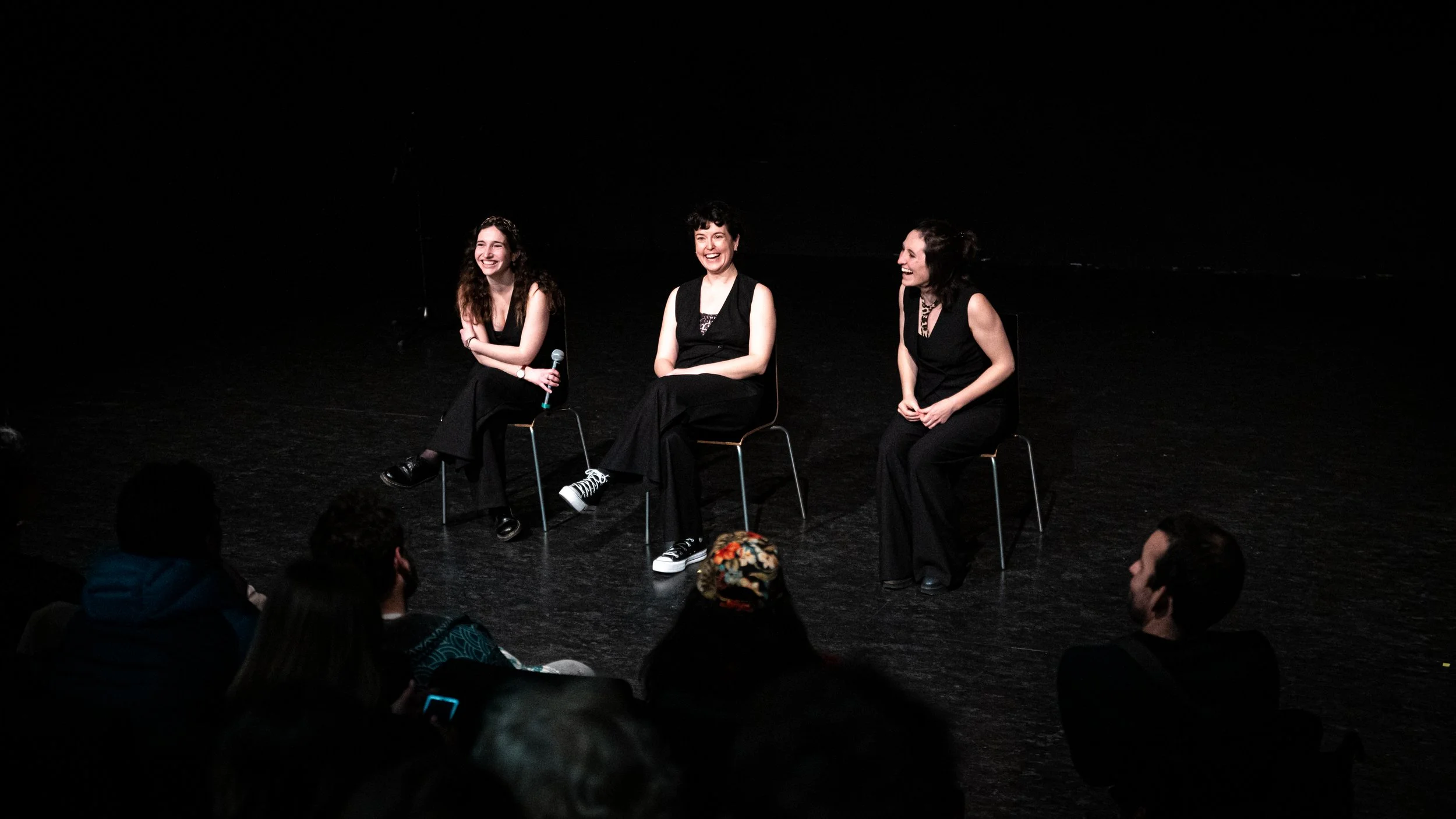 Trois femmes assises sur scène en noir, partageant un moment de rire, face à un public.
