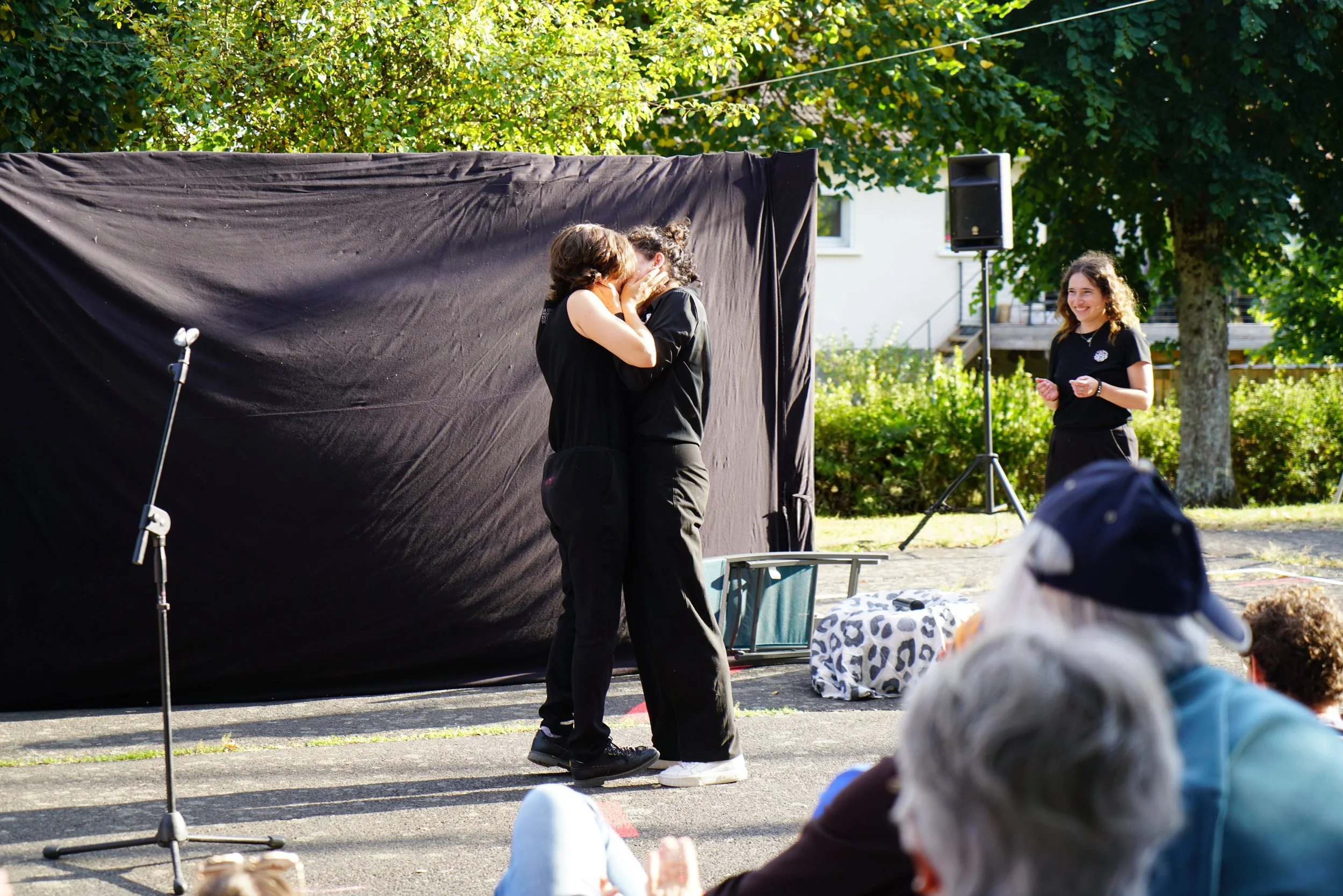 Deux personnes s'embrassent dans un spectacle en plein air avec un fond noir, alors qu'une autre personne regarde et sourit. Il y a des spectateurs assis en face, des arbres verts et une enceinte visible en arrière-plan.