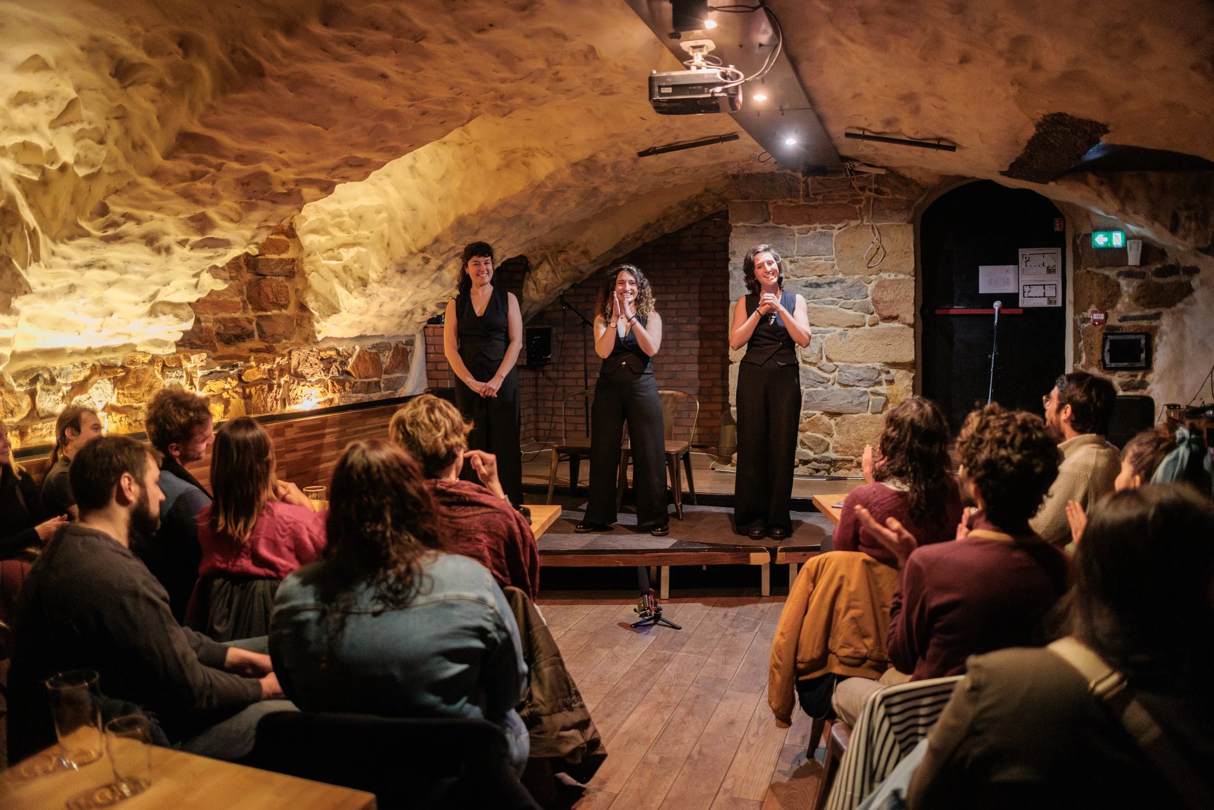 Trois femmes en tenue noire se produisent sur scène devant une audience dans une salle avec un plafond en voûte en pierre, un éclairage chaleureux et un décor en brique et pierre.
