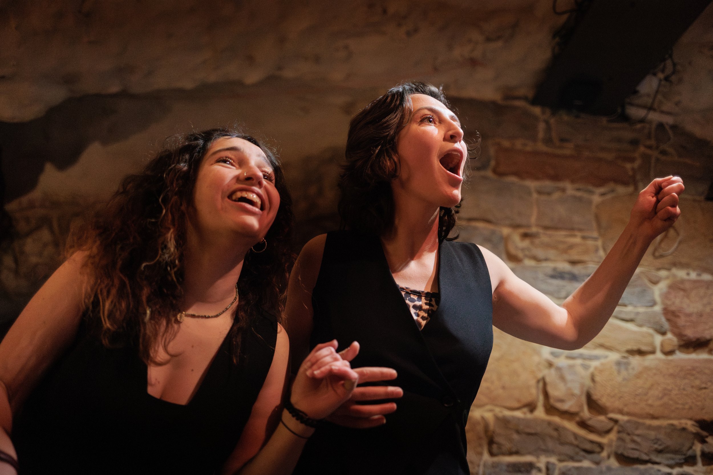 Deux femmes, l'une brune aux cheveux bouclés et l'autre à cheveux courts, souriantes, parlent ou chantent ensemble dans une pièce avec un mur en pierre, créant une atmosphère chaleureuse et conviviale.