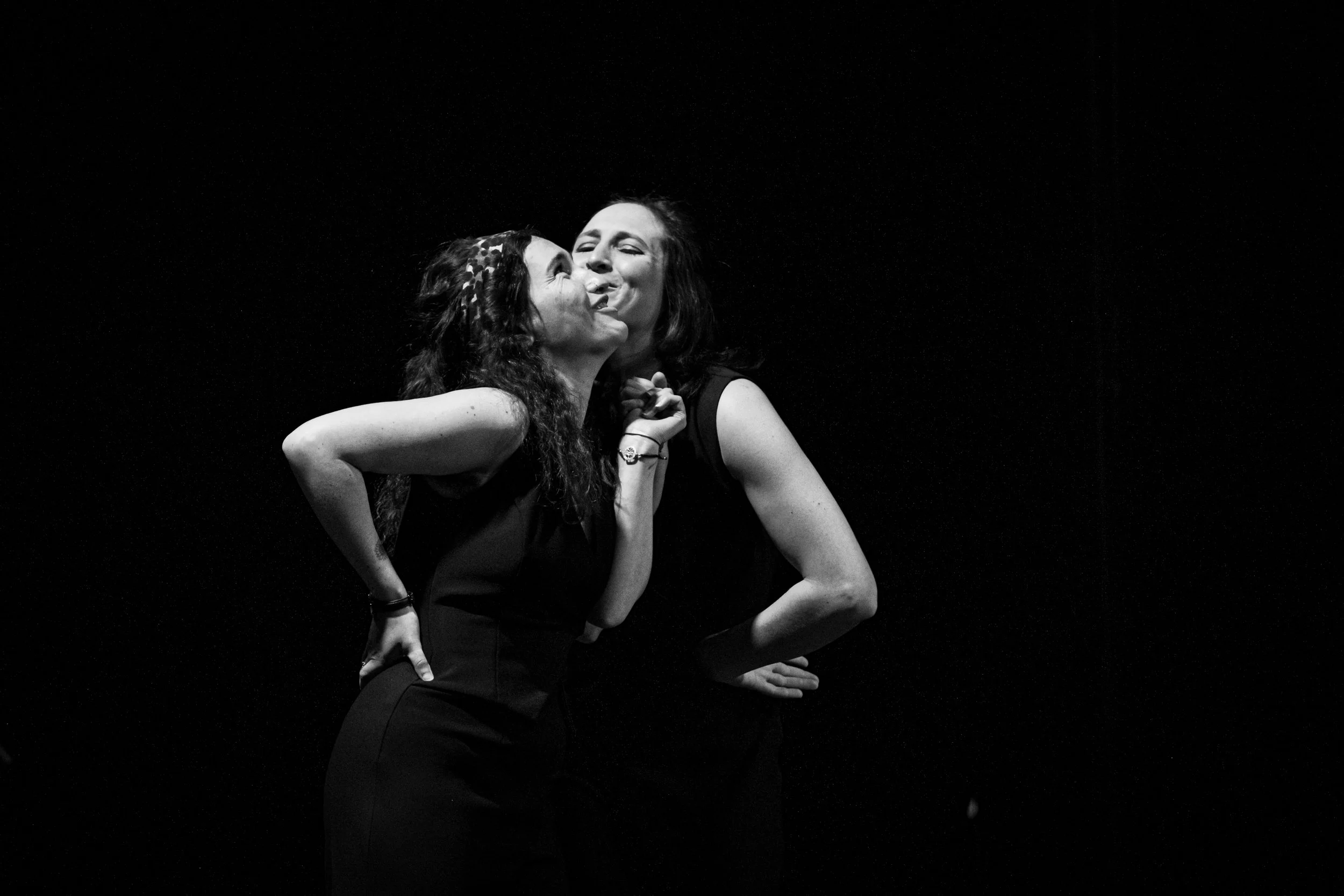 Deux femmes en noir et blanc, une embrassant l'autre sur la joue, dans une pose expressive et intime, contre un fond noir.