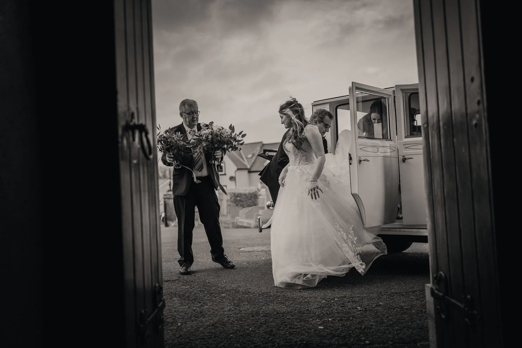 A bride getting out of a vintage car with the help of her groom and an older man holding a bouquet of flowers, seen through an open door frame in black and white.