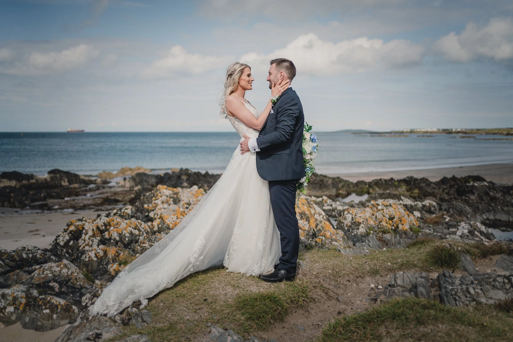 A bride and groom embrace on a rocky beach during their wedding, with the ocean and partly cloudy sky in the background.