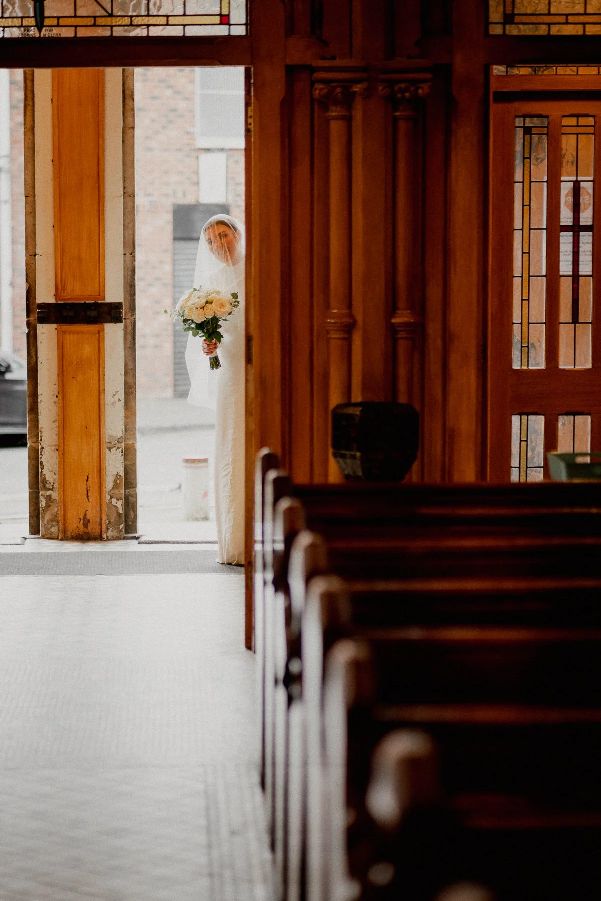 A bride holding a bouquet of white roses, standing in a doorway and looking inside a church.