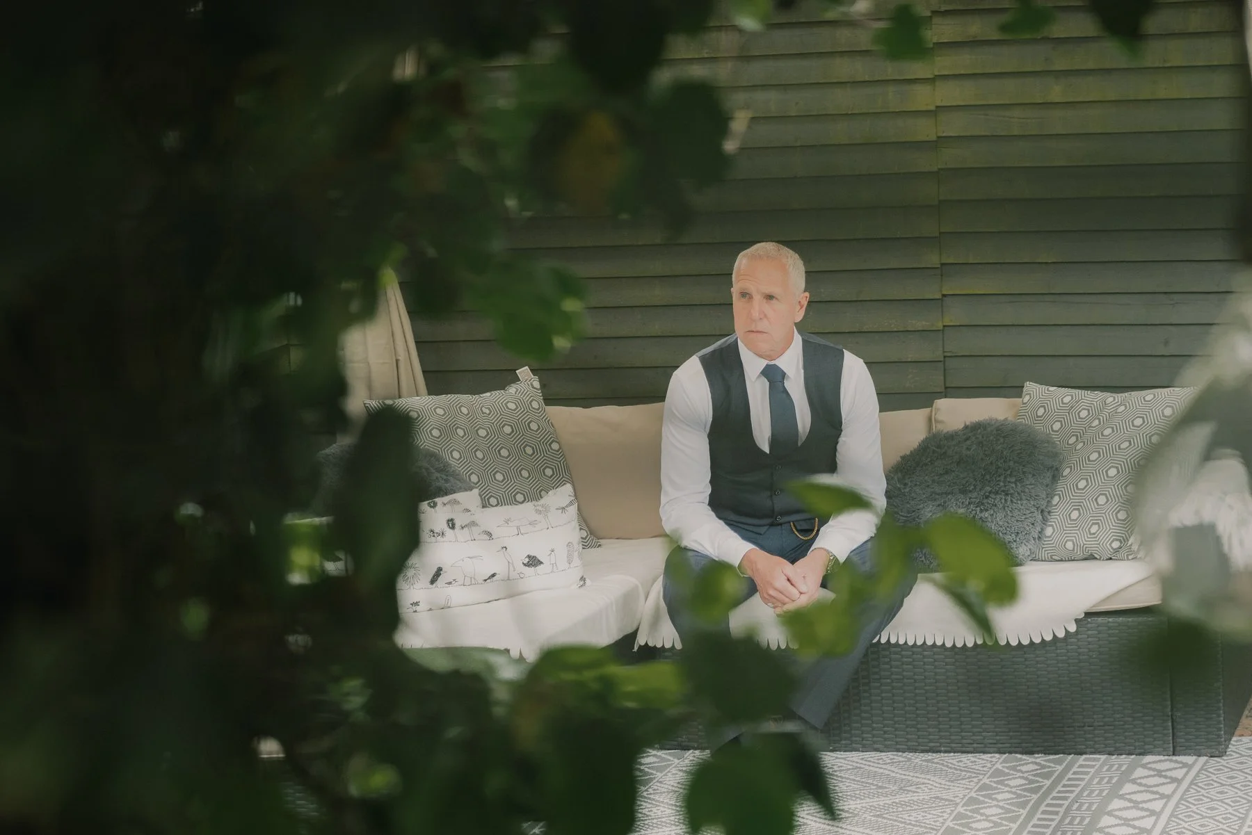 A man in a black vest and tie sitting on a beige outdoor sofa, visible through green foliage, with pillows against a dark green wooden wall.