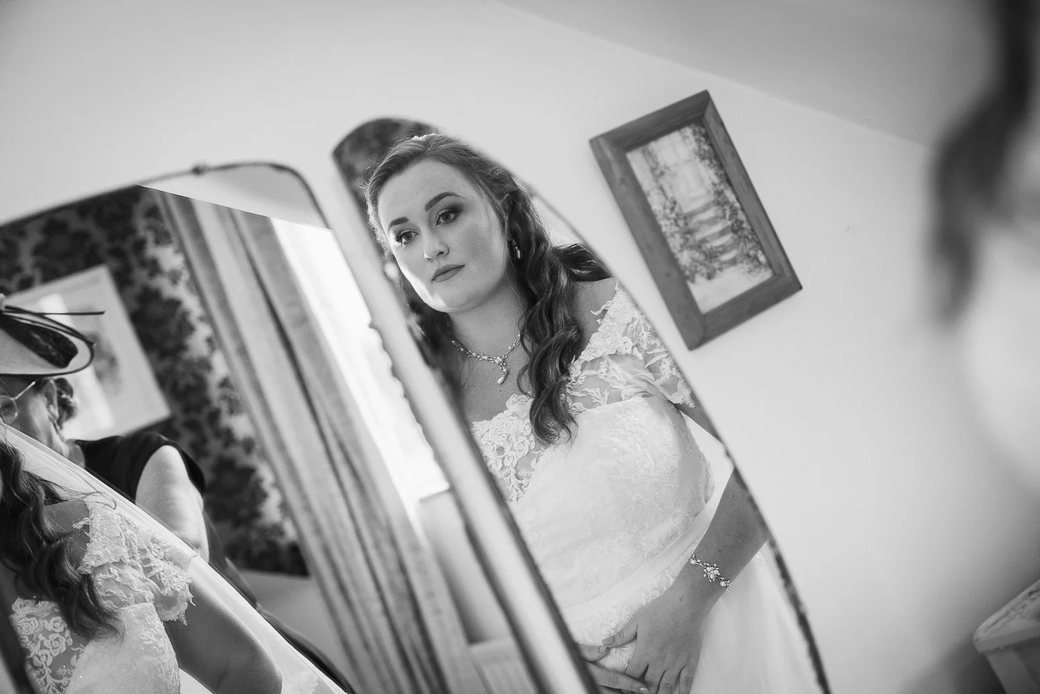 A woman in a wedding dress looking at herself in a mirror, with her hair styled in loose curls, wearing jewelry. The photograph is in black and white.