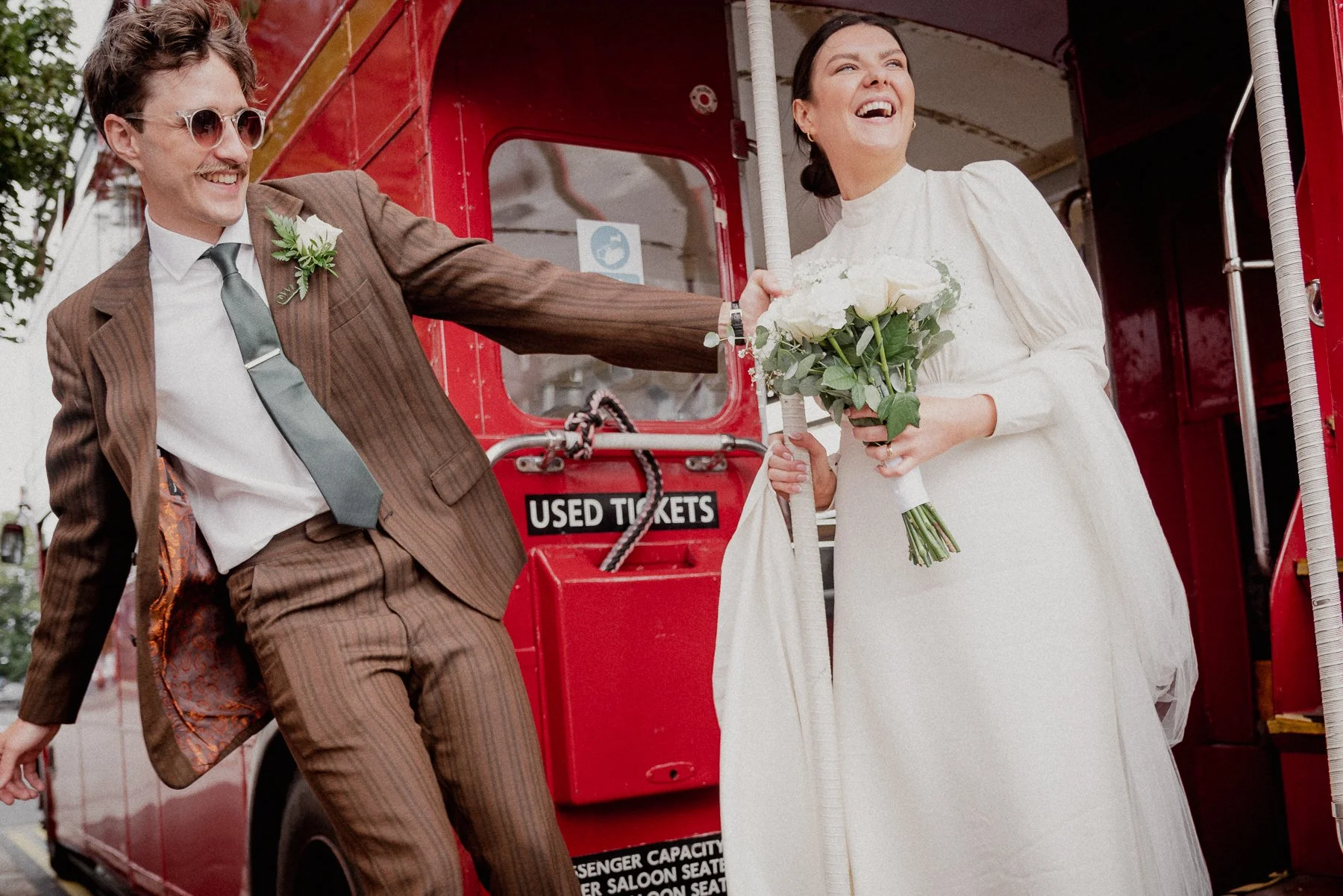 A happy bride and groom standing next to a red ticket booth, with the groom playfully pulling on the bride's wedding dress as they smile.