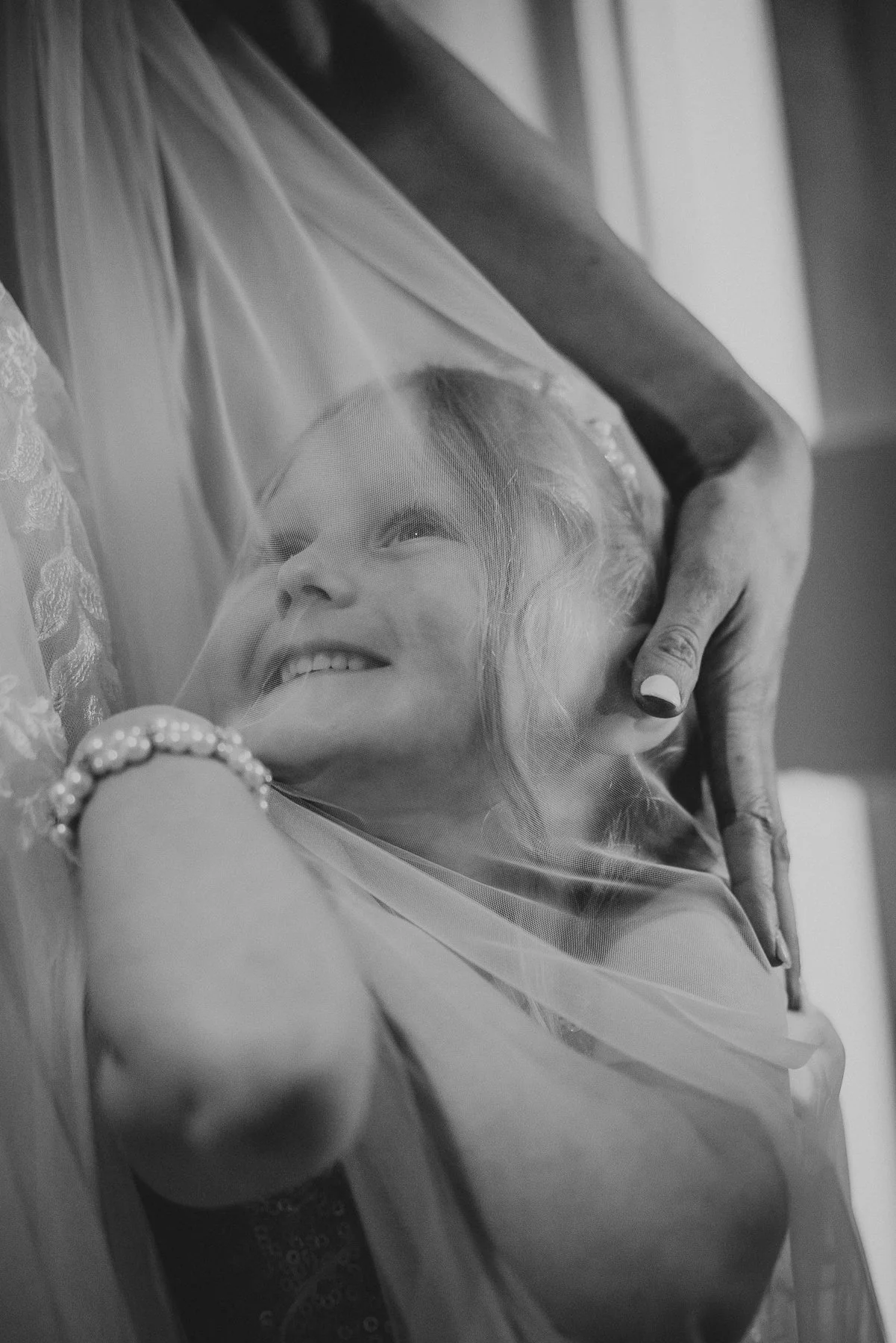 A joyful elderly woman smiling while lying down with a sheer veil over her face, being gently held by someone with well-manicured nails and a bracelet.