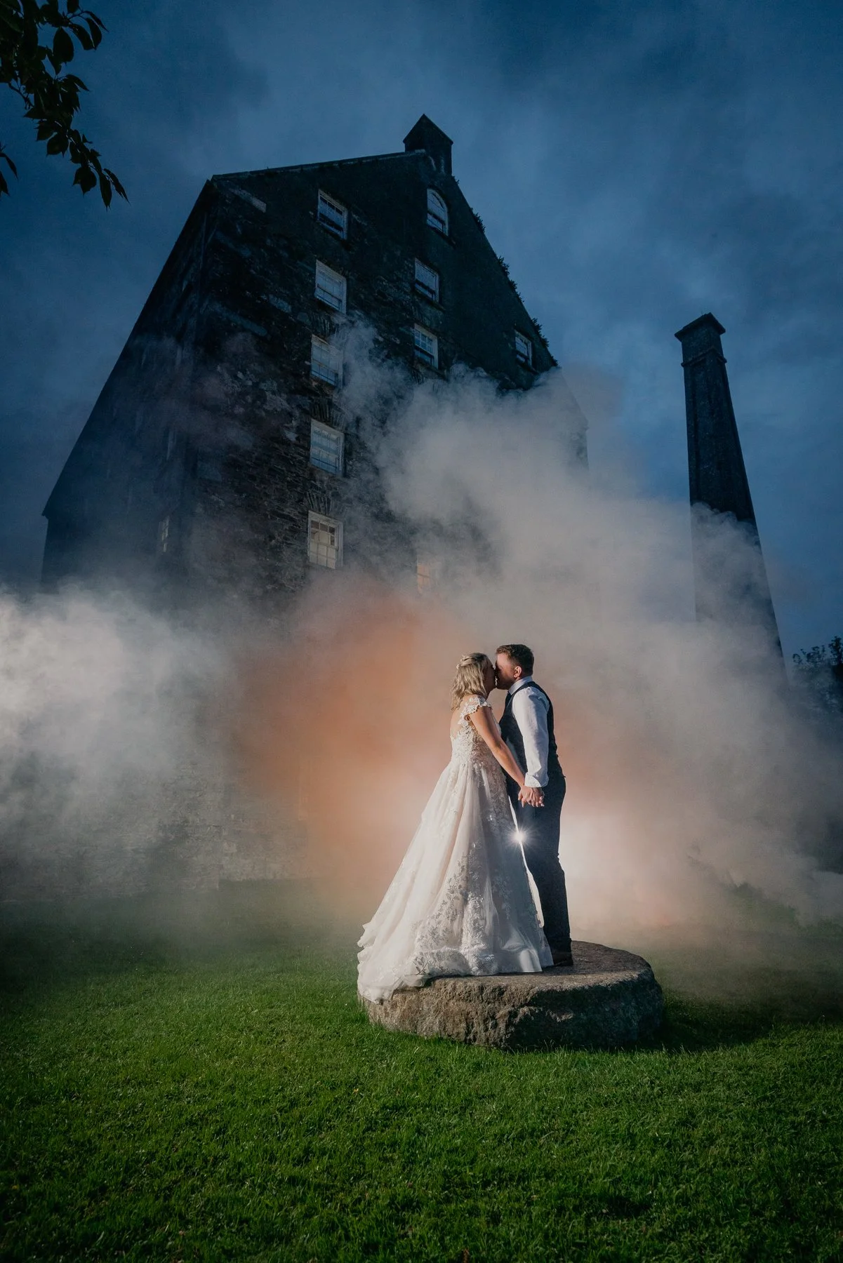 A bride and groom kissing on a stone platform outdoors, with an old brick building and chimney in the background, surrounded by mist in a moody, romantic setting at dusk or night.