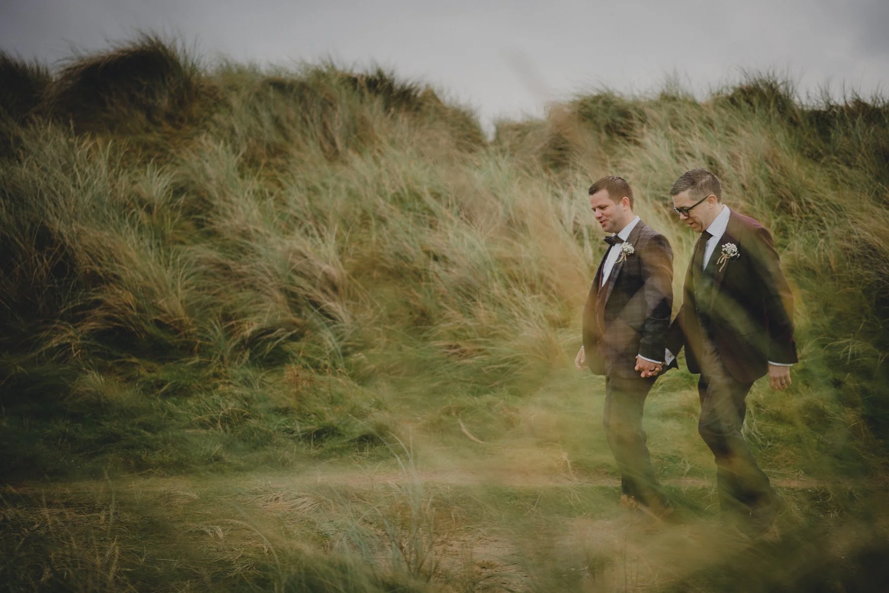Two men dressed in suits and bow ties, holding hands and walking through tall green grass on a cloudy day.