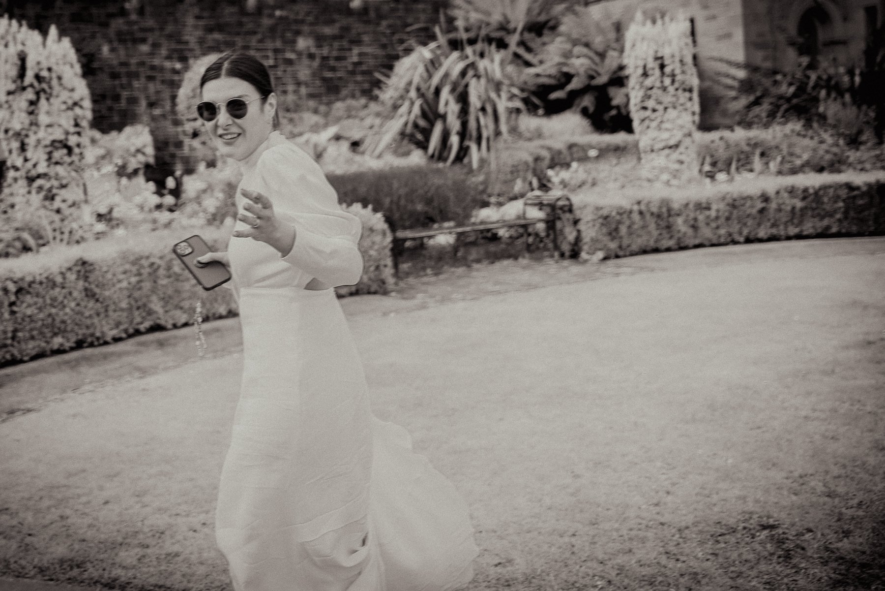 A woman wearing sunglasses and a long white dress is smiling and waving at the camera. She is holding a phone in her right hand. The background features a garden with plants, bushes, and a brick building.