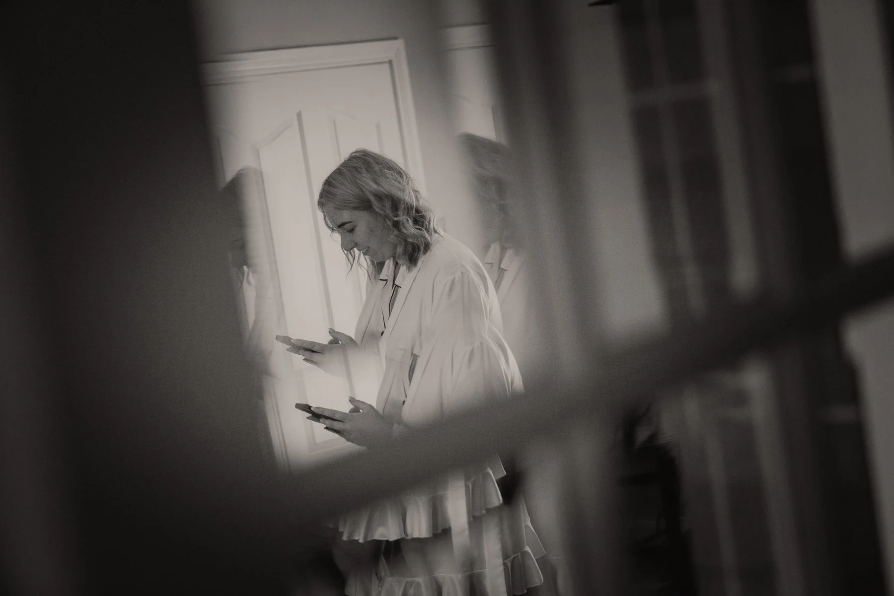 A woman in a white dress looking at her phone, seen through an opening in a wall or piece of furniture, creating a blurred frame around her.