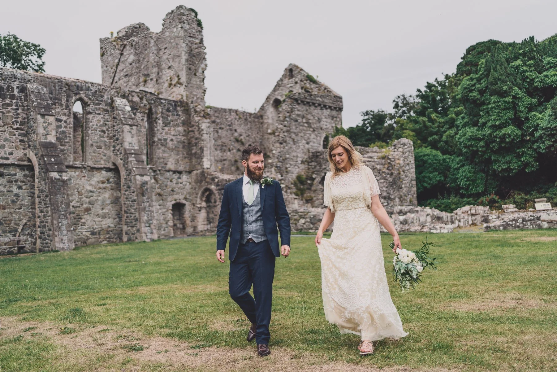 A couple in wedding attire walking on grass in front of an old stone ruins with green trees in the background.
