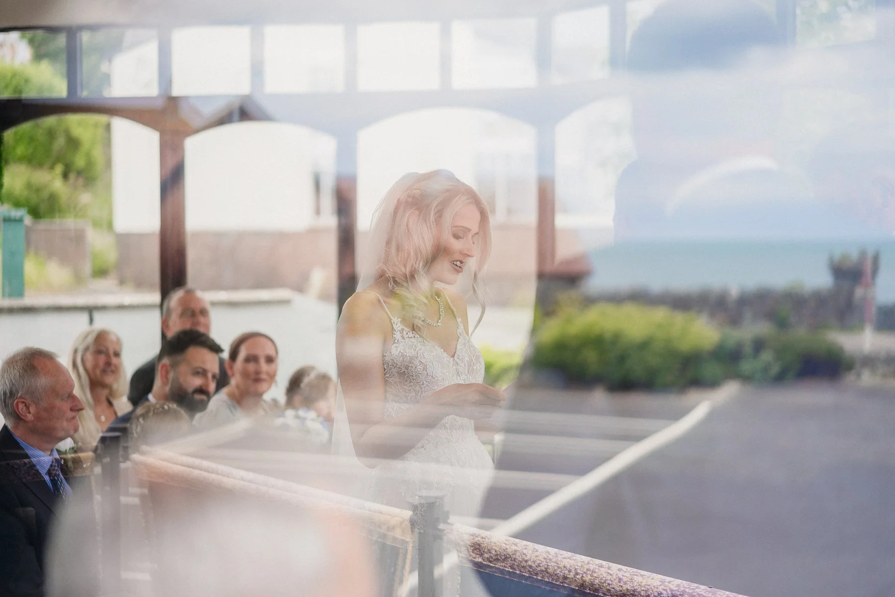 A bride reading a speech during a wedding reception, reflected in a window, with guests sitting at a table behind her.