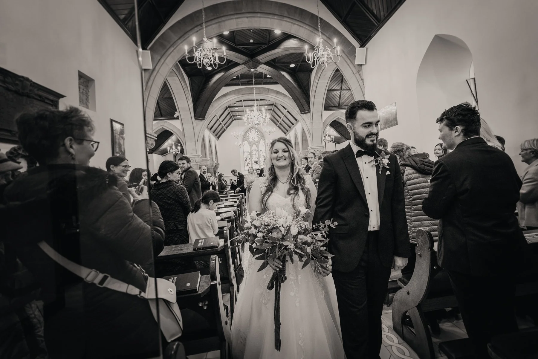 A black and white photo of a wedding ceremony inside a church. The bride, wearing a white wedding dress and holding a bouquet of flowers, is smiling as she walks down the aisle with the groom, who is dressed in a tuxedo, both surrounded by seated gue