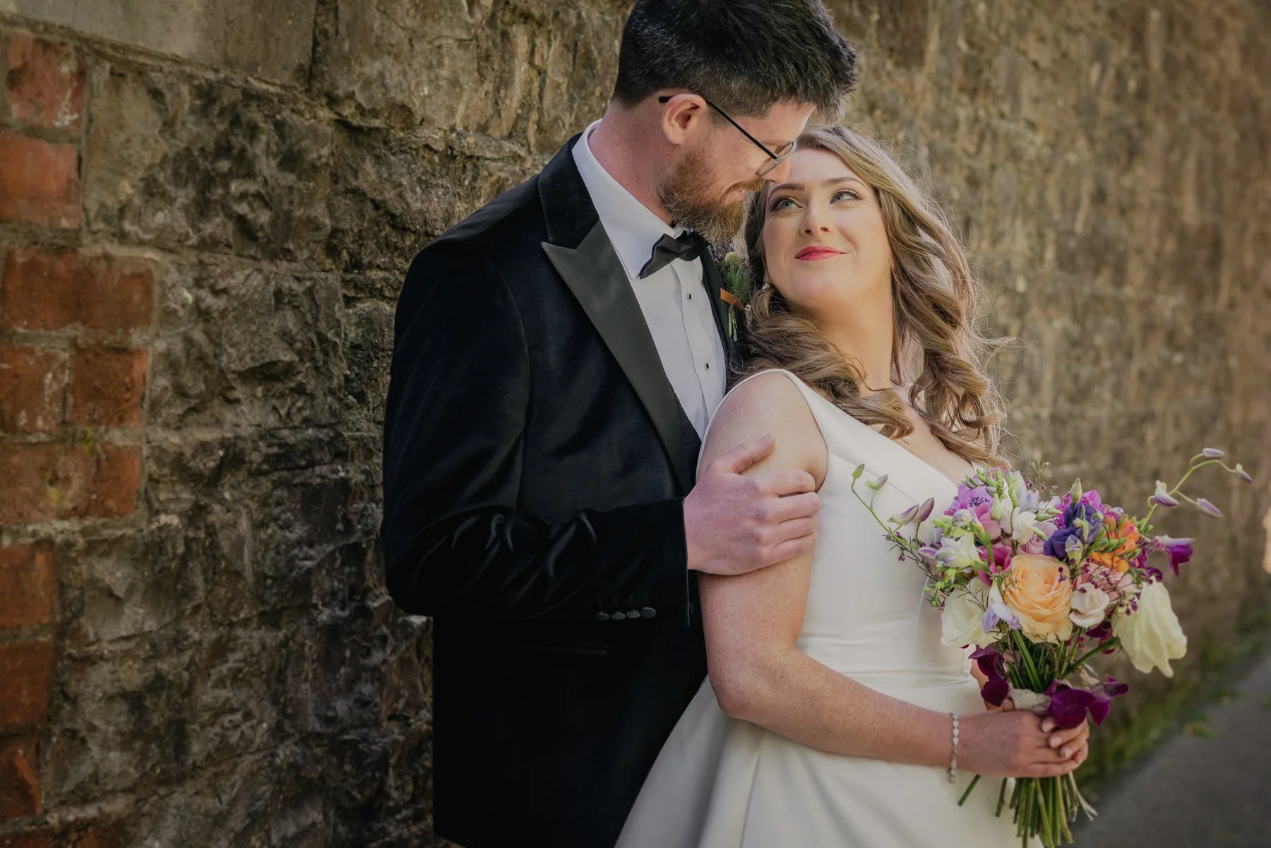 A bride and groom standing close together, leaning their foreheads against each other, in front of a brick wall. The bride is holding a large bouquet of colorful flowers, and the groom is dressed in a black tuxedo with a bow tie.