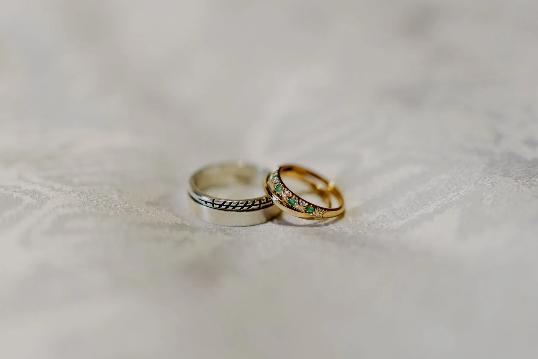 Two rings, one silver with engraved design and one gold with colorful gemstones, resting on a soft white fabric.