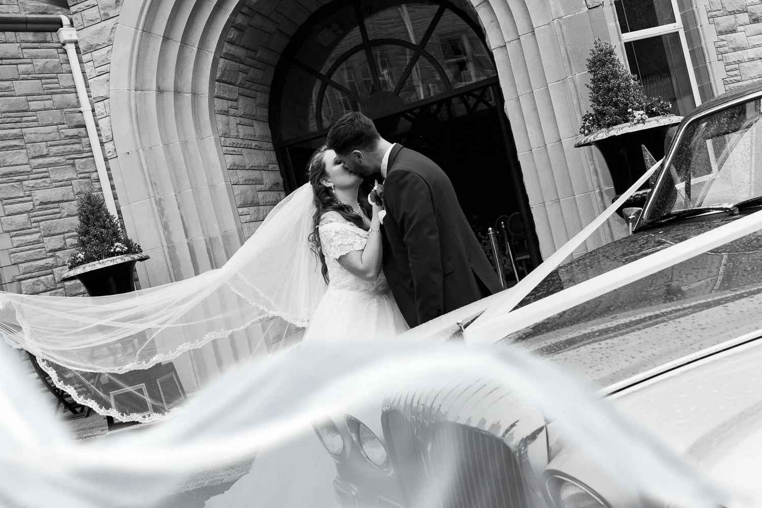 A black and white photo of a bride and groom kissing in front of a building, with a vintage car decorated with ribbons in the foreground.