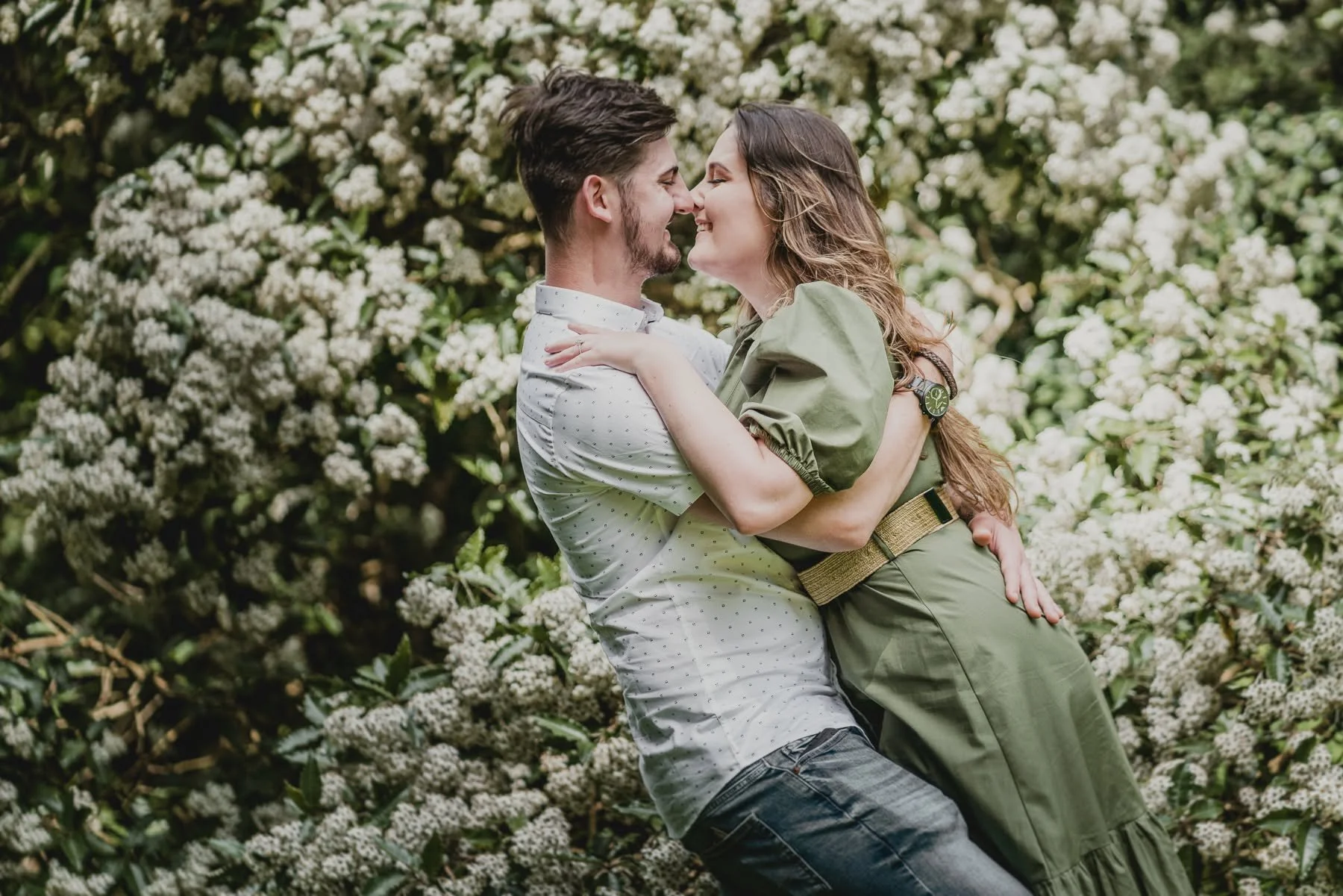A couple is embracing outdoors in front of white flowering bushes, smiling and looking at each other close to their noses.
