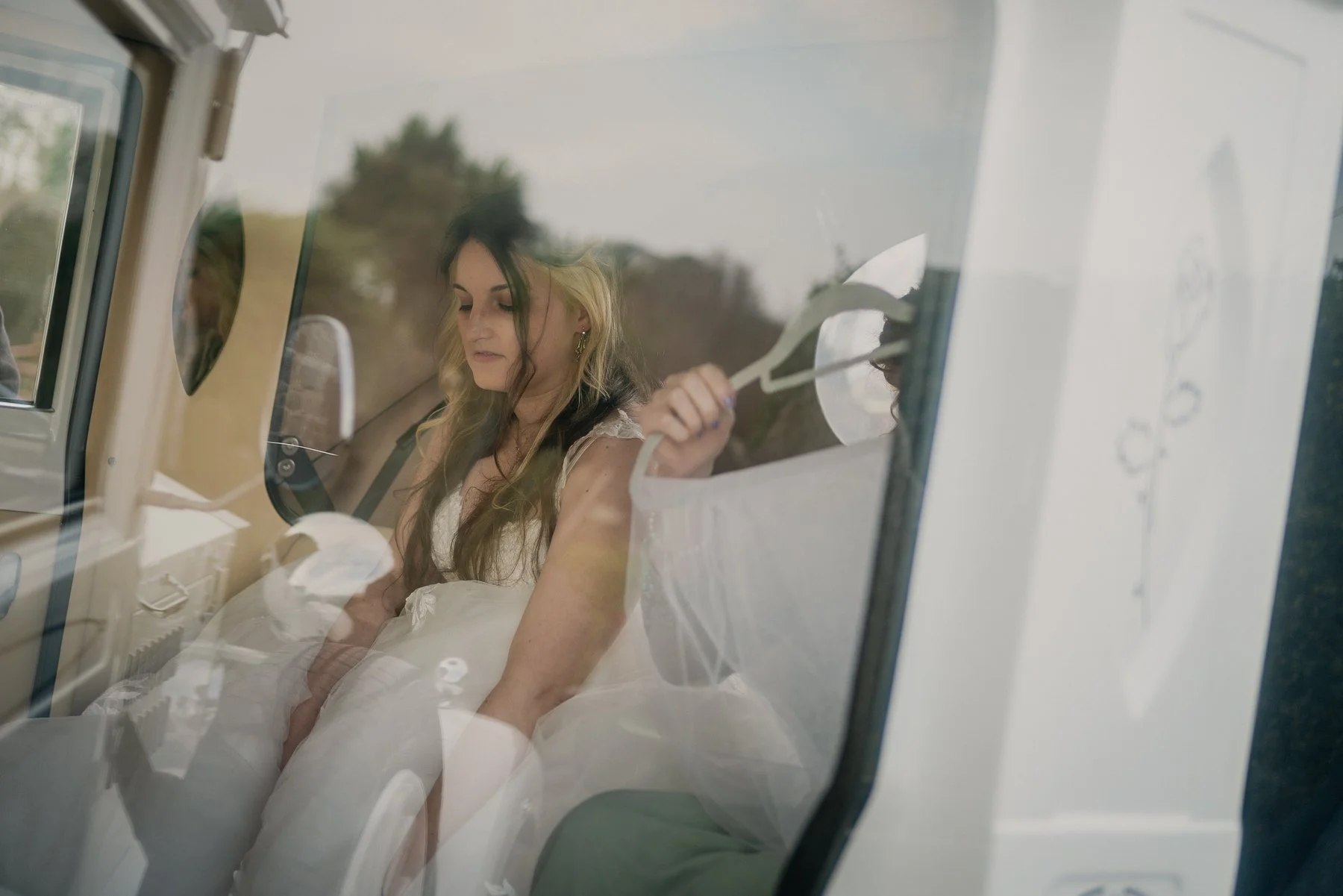 A young woman with wavy hair and earrings, wearing a white dress, sitting inside a vintage vehicle, holding a white rose, with trees and cloudy sky visible through the window.